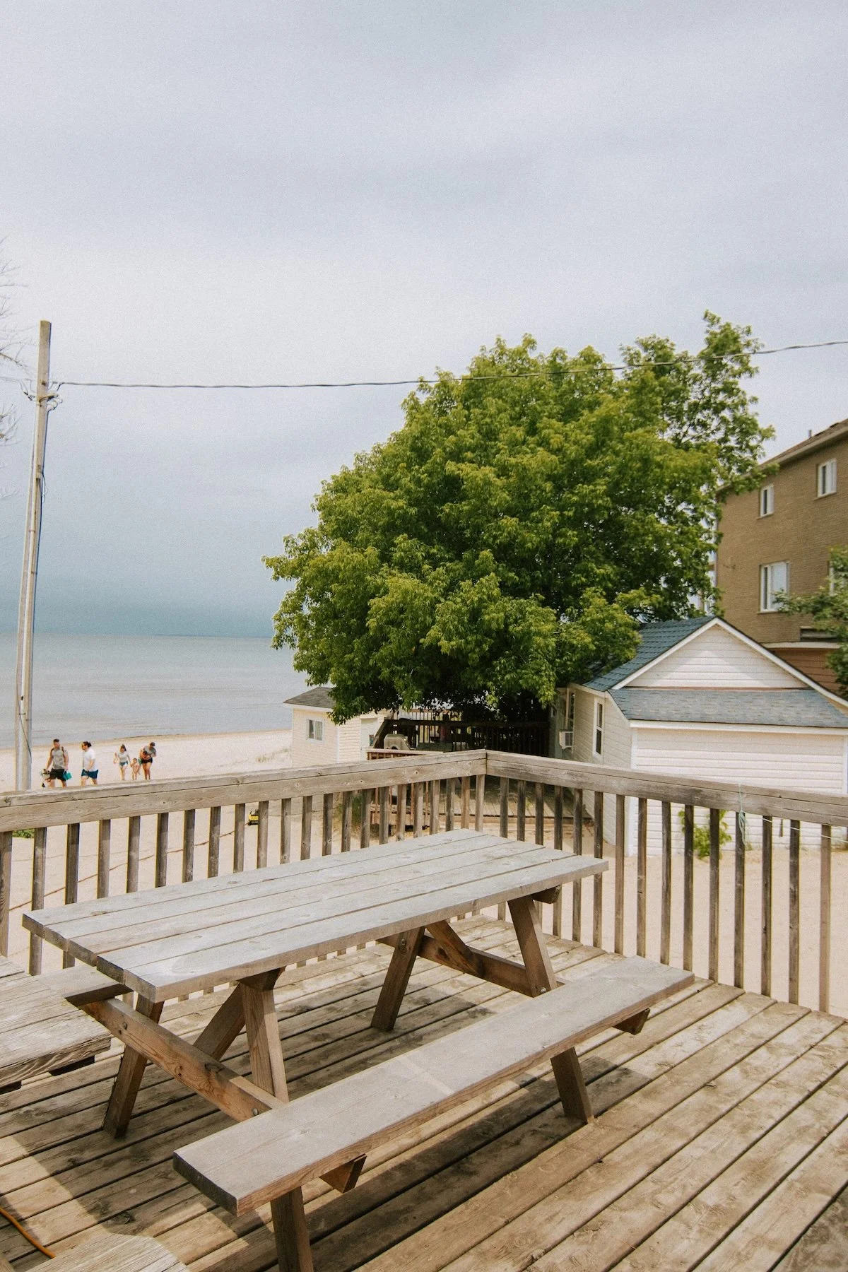 Wooden picnic tables on a beachfront deck overlooking a sandy beach, lake, trees, and nearby cottages