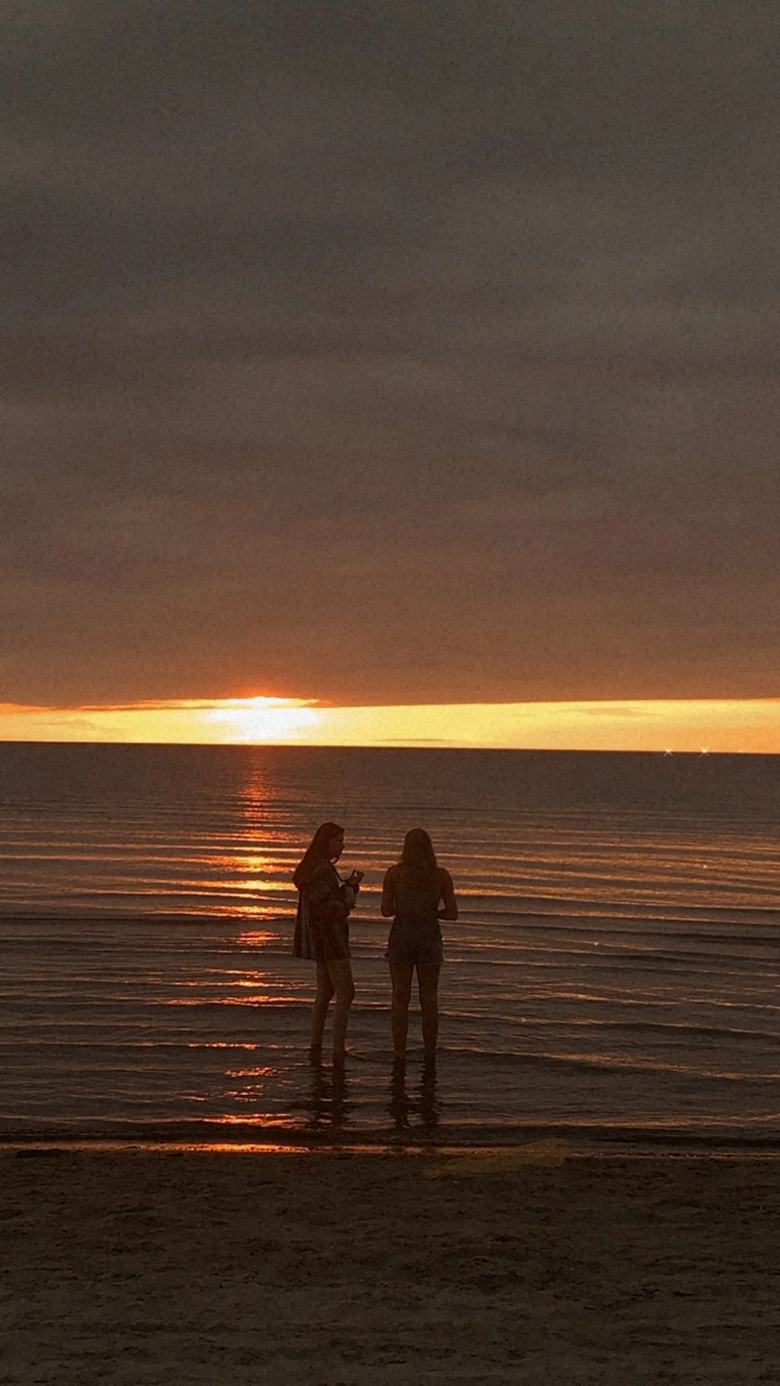 Two silhouetted people standing in shallow ocean water at sunset, with golden reflections across the waves.