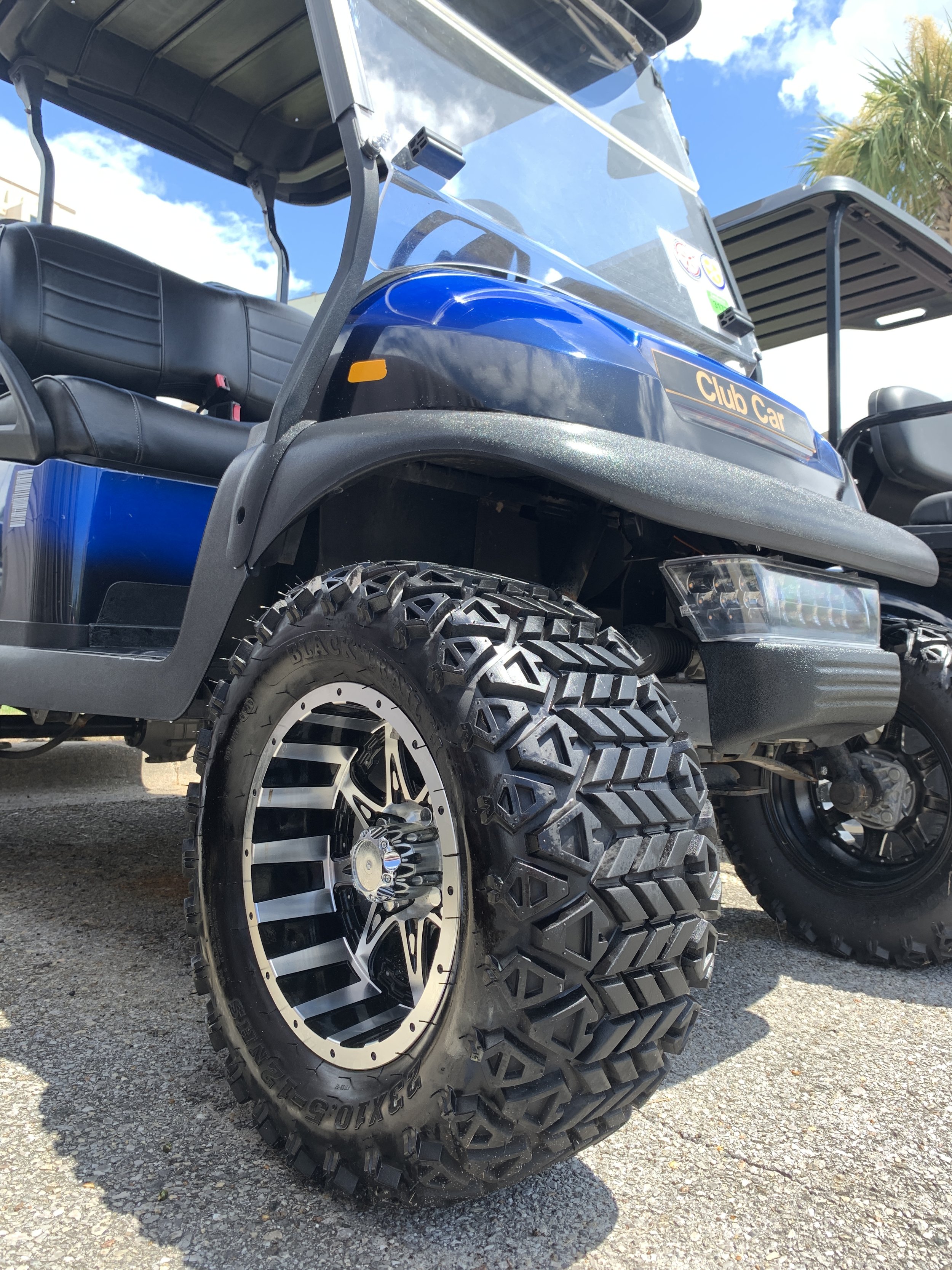 Close-up of a blue lifted Club Car golf cart with oversized off-road tires, custom alloy wheels, windshield, and black seats.