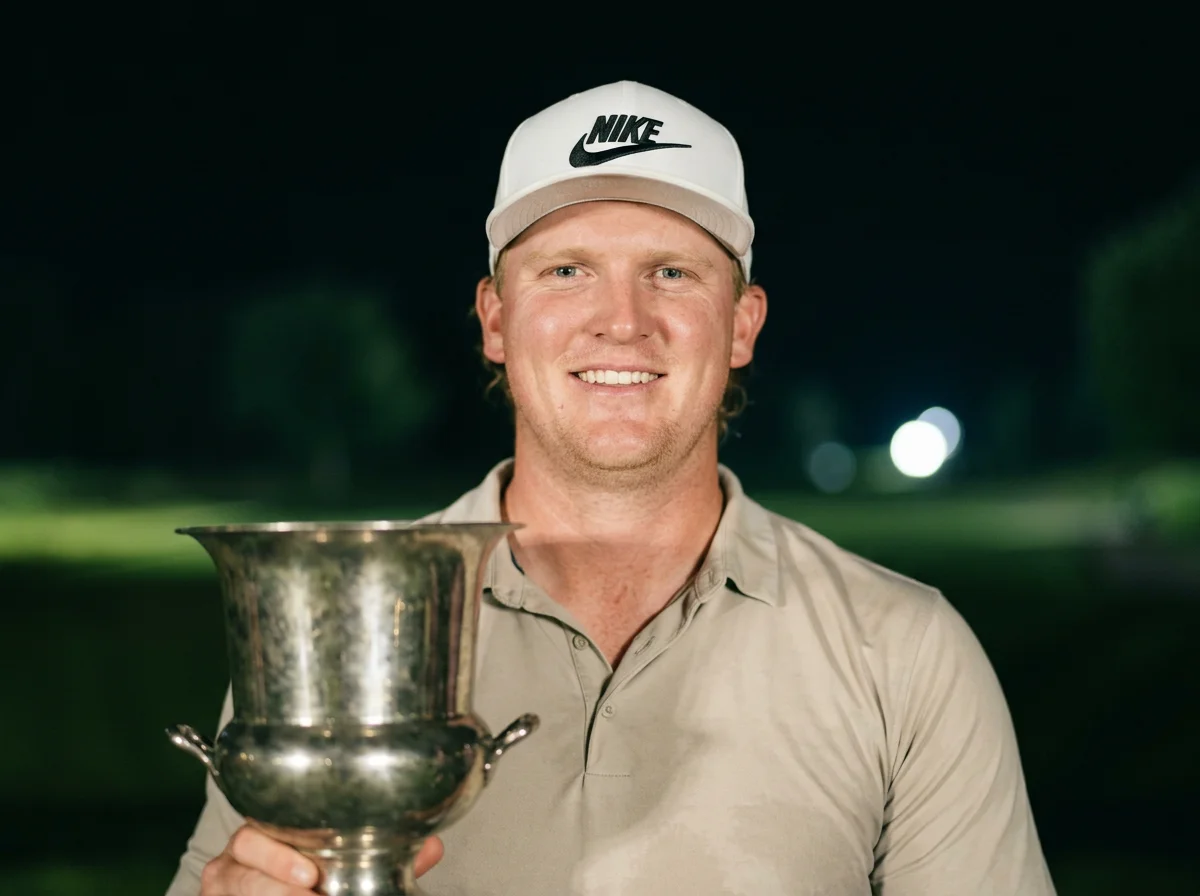Smiling man wearing a Nike cap and holding a large silver trophy outdoors at night