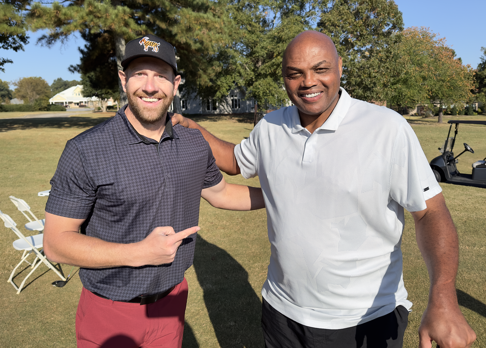 Two smiling men pose on a golf course, with one pointing toward the other beside a golf cart. Josh Cassaday and Charles Barkley