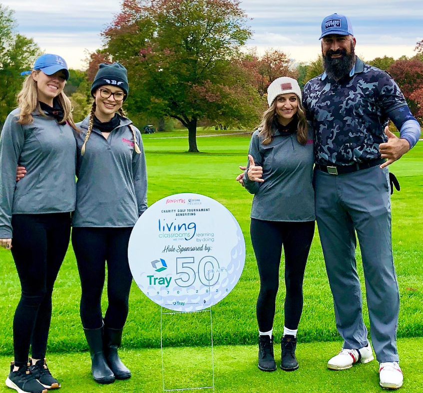 Four people pose on a golf course beside a sign for the Charity Golf Tournament benefiting Living Classrooms and celebrating Tray’s 50th anniversary.