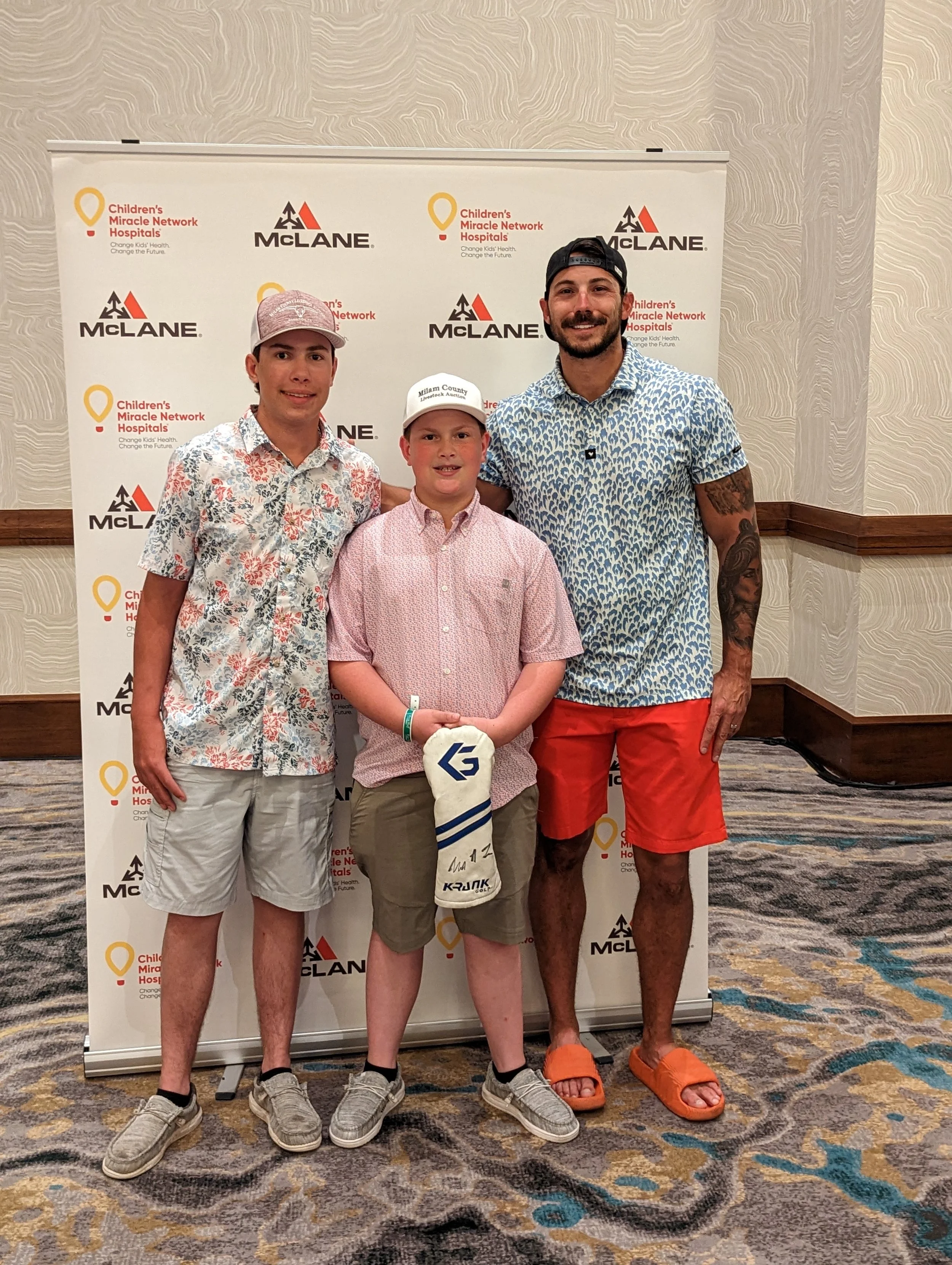 Three people pose in front of a Children's Miracle Network Hospitals and McLane event backdrop, with a child holding a signed golf club headcover.