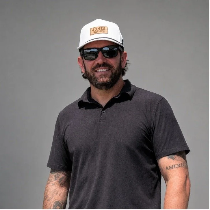 Bearded man wearing sunglasses, a white Jones cap, and black polo shirt against a gray background
