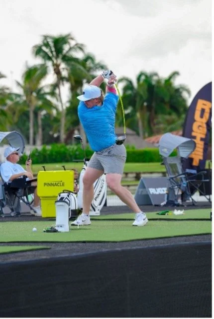 Golfer in a blue shirt swinging a club at an outdoor driving range with palm trees