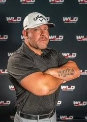 Man wearing a Callaway golf cap and black polo shirt with a tattooed forearm, standing before a WLD-branded backdrop