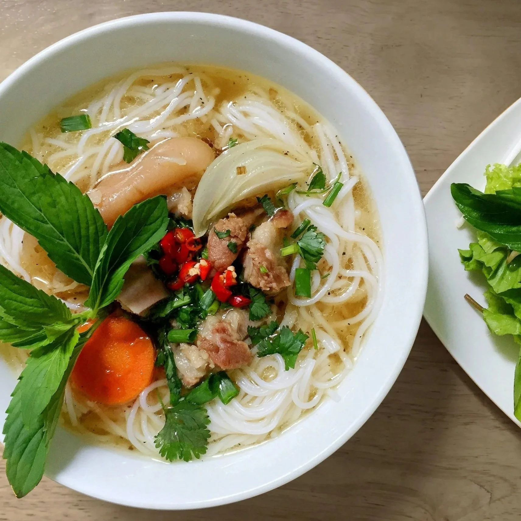 Bowl of Vietnamese rice noodle soup with pork, sausage, onions, herbs, carrots, and red chili peppers, served with fresh greens