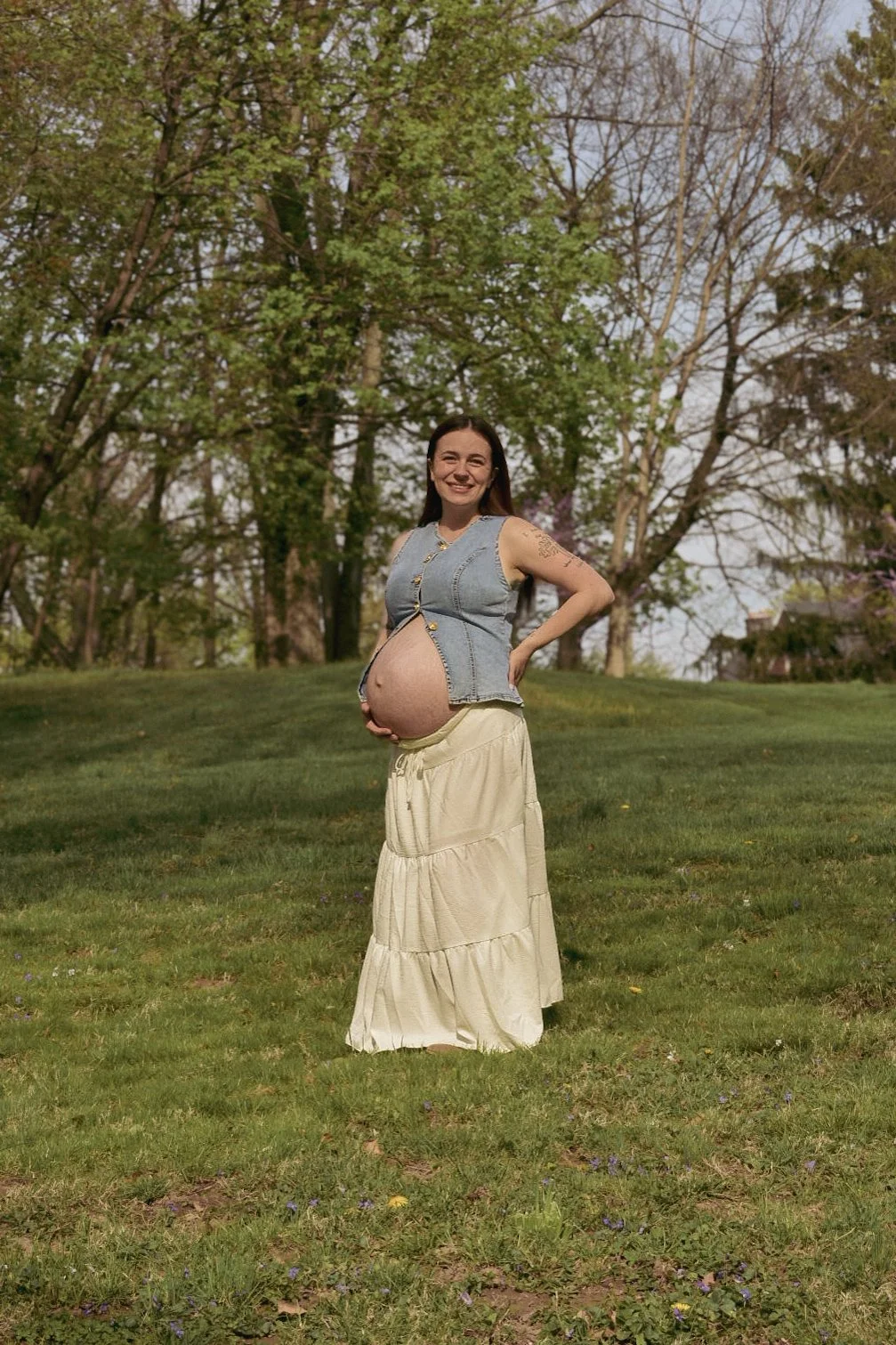 Pregnant woman posing in a grassy park, wearing a denim vest and long cream skirt.