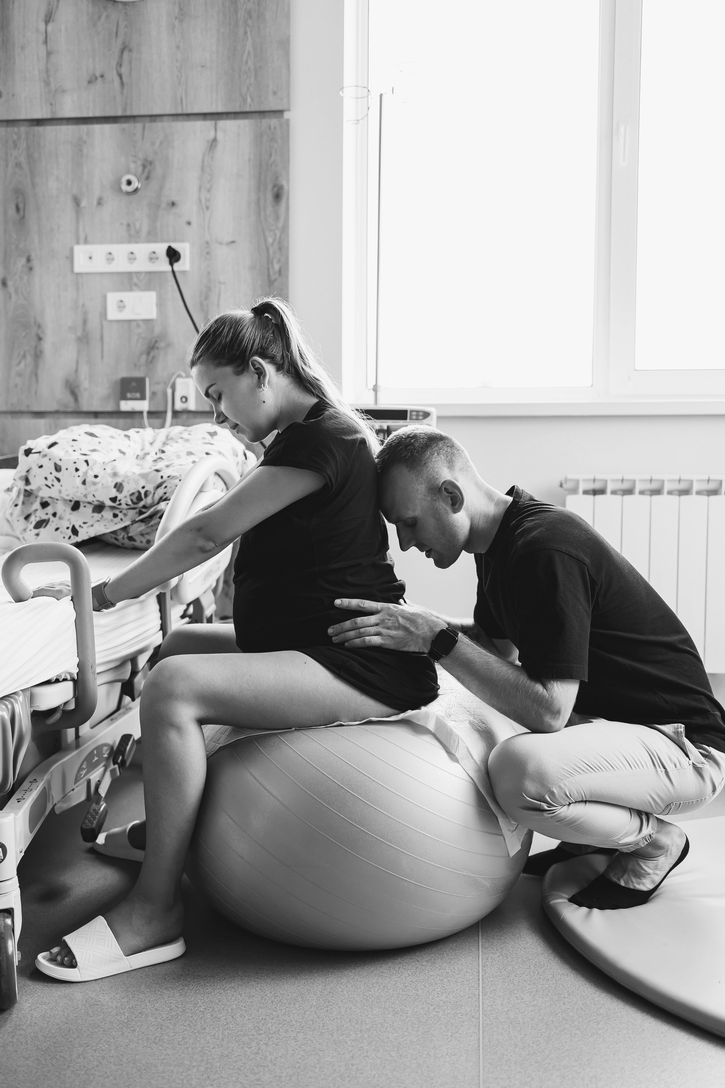 Pregnant woman laboring on a birthing ball in a hospital room while a partner provides support.