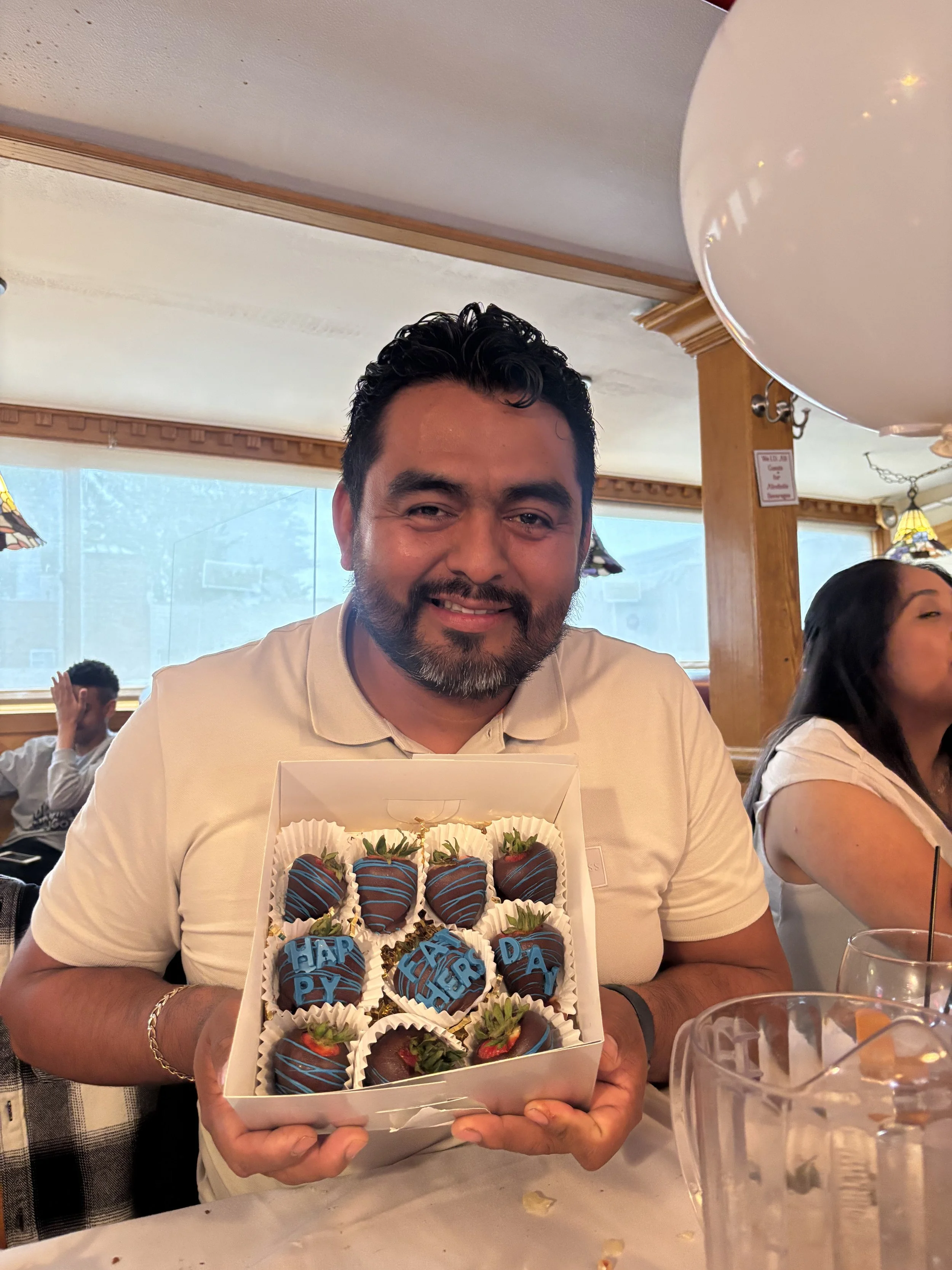 Smiling man holding a box of chocolate-covered strawberries decorated with blue drizzle and lettering at a restaurant table