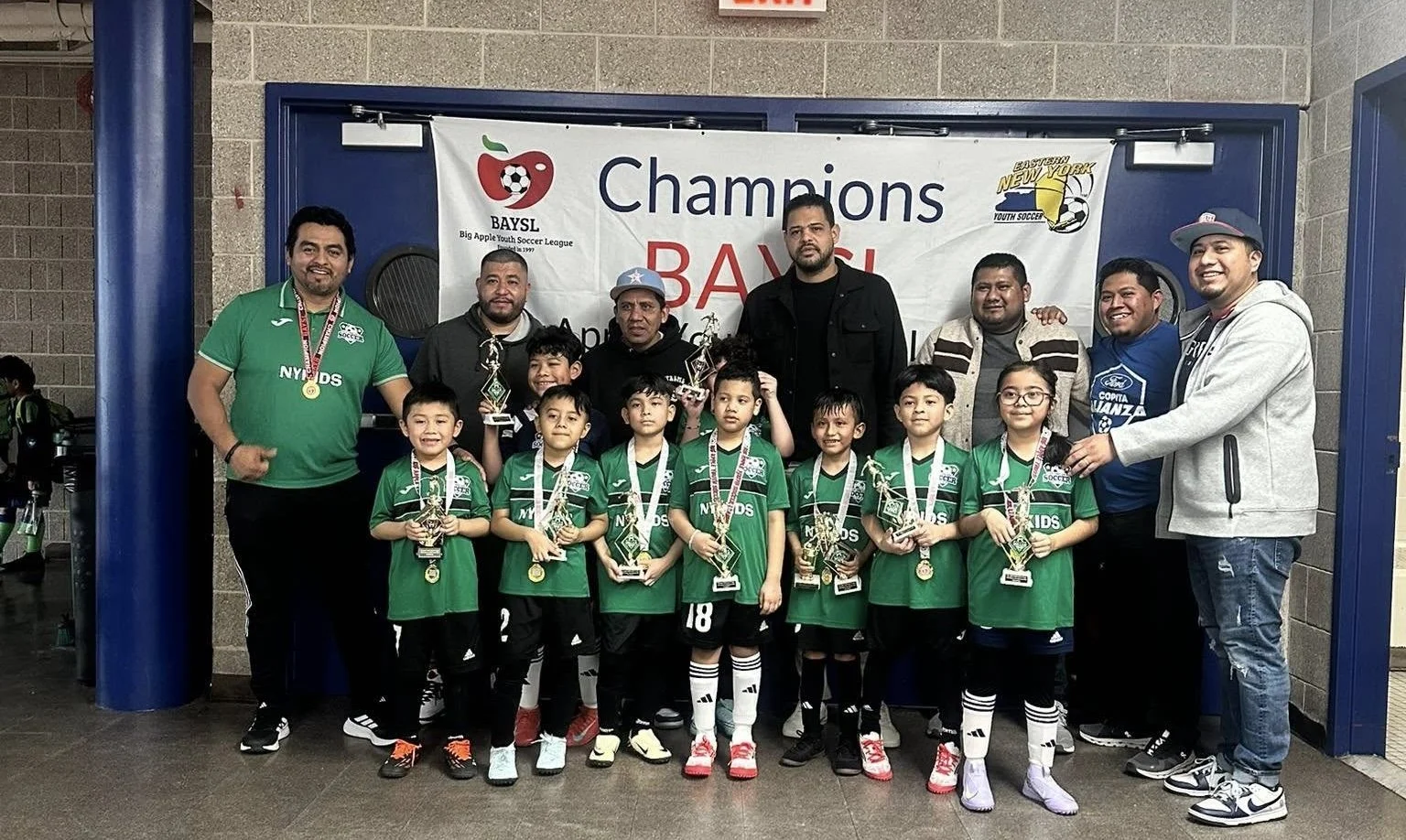Youth soccer team in green uniforms posing with coaches and trophies beneath a BAYSL champions banner