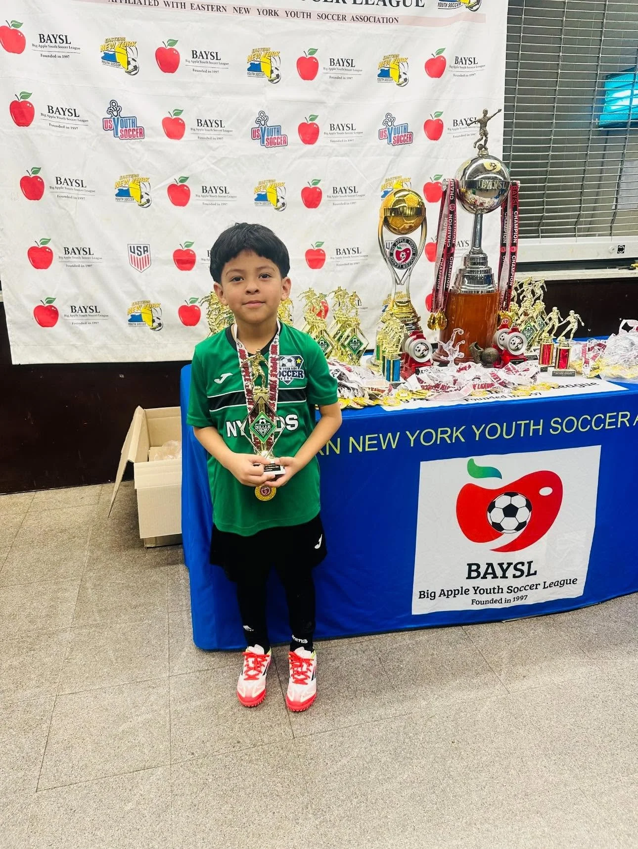 Young soccer player holding a trophy and medal beside a youth soccer awards table with cups and medals