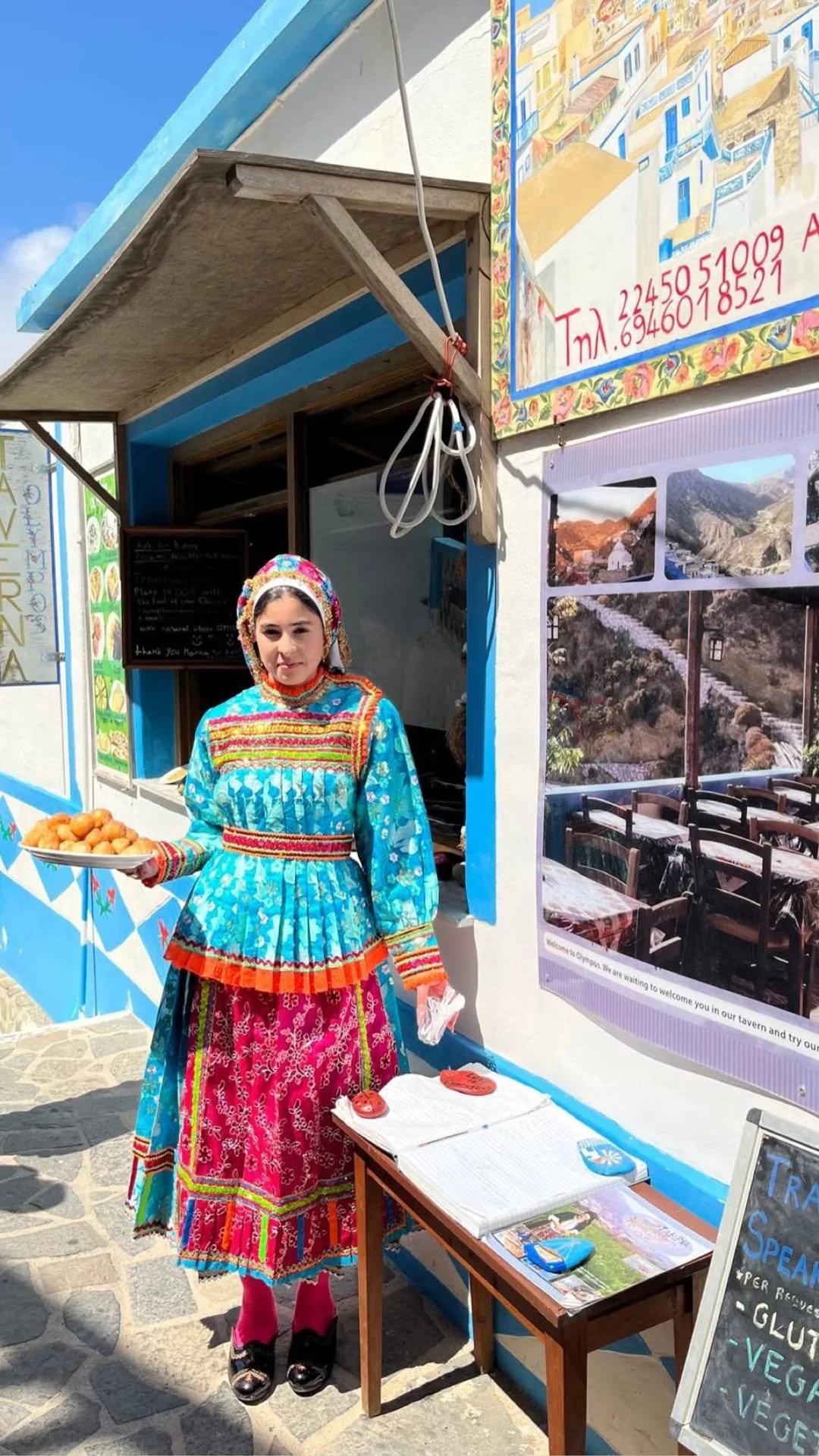 Woman in colorful traditional Greek clothing holding a tray of pastries outside a taverna in Greece