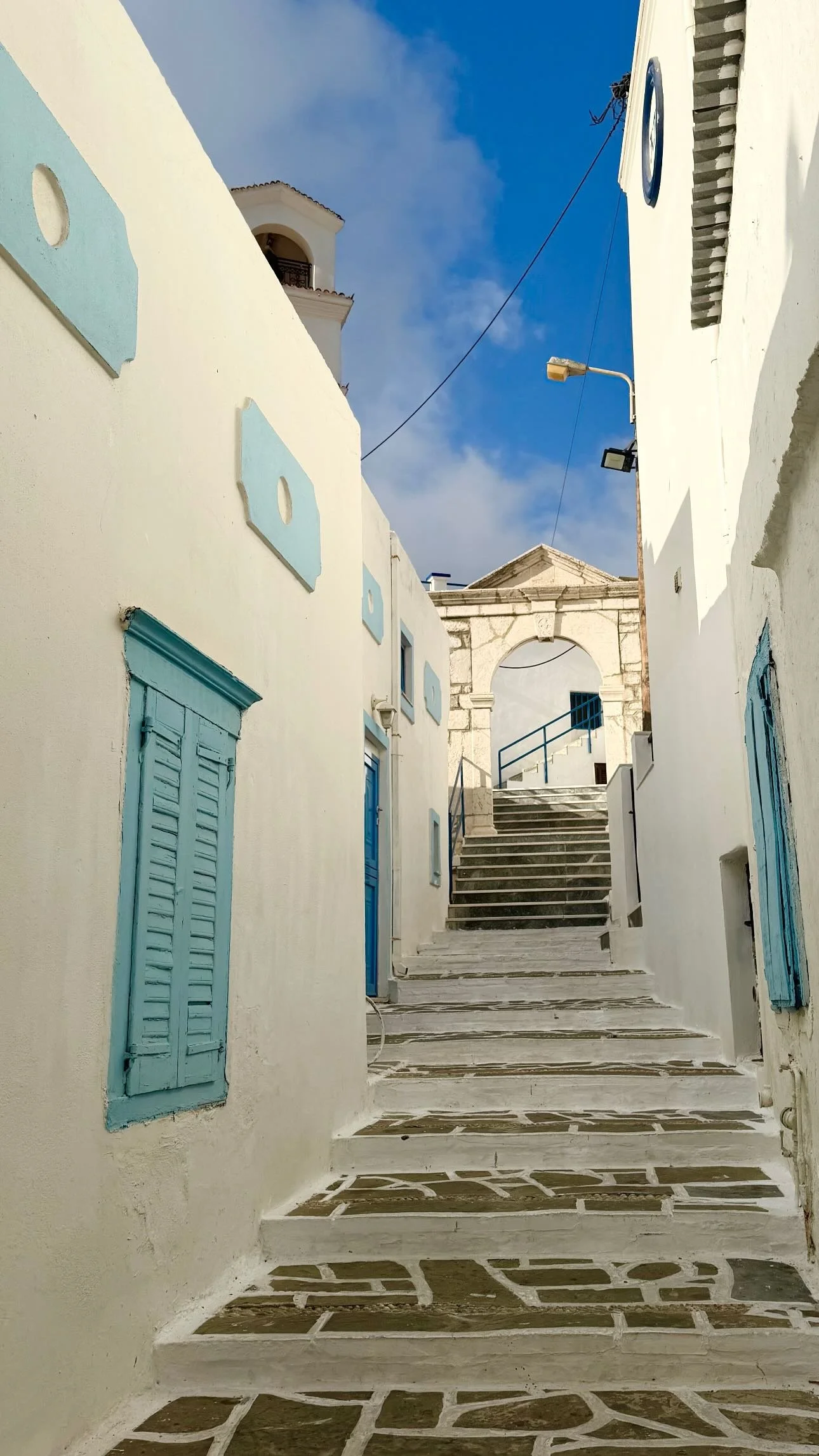 Narrow whitewashed Greek village alley with turquoise shutters, stone steps, and an arched gateway under a blue sky