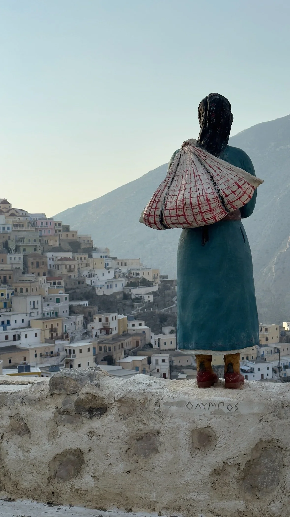 Statue of a traditional Greek woman carrying a large bundle overlooking a hillside village and mountains.