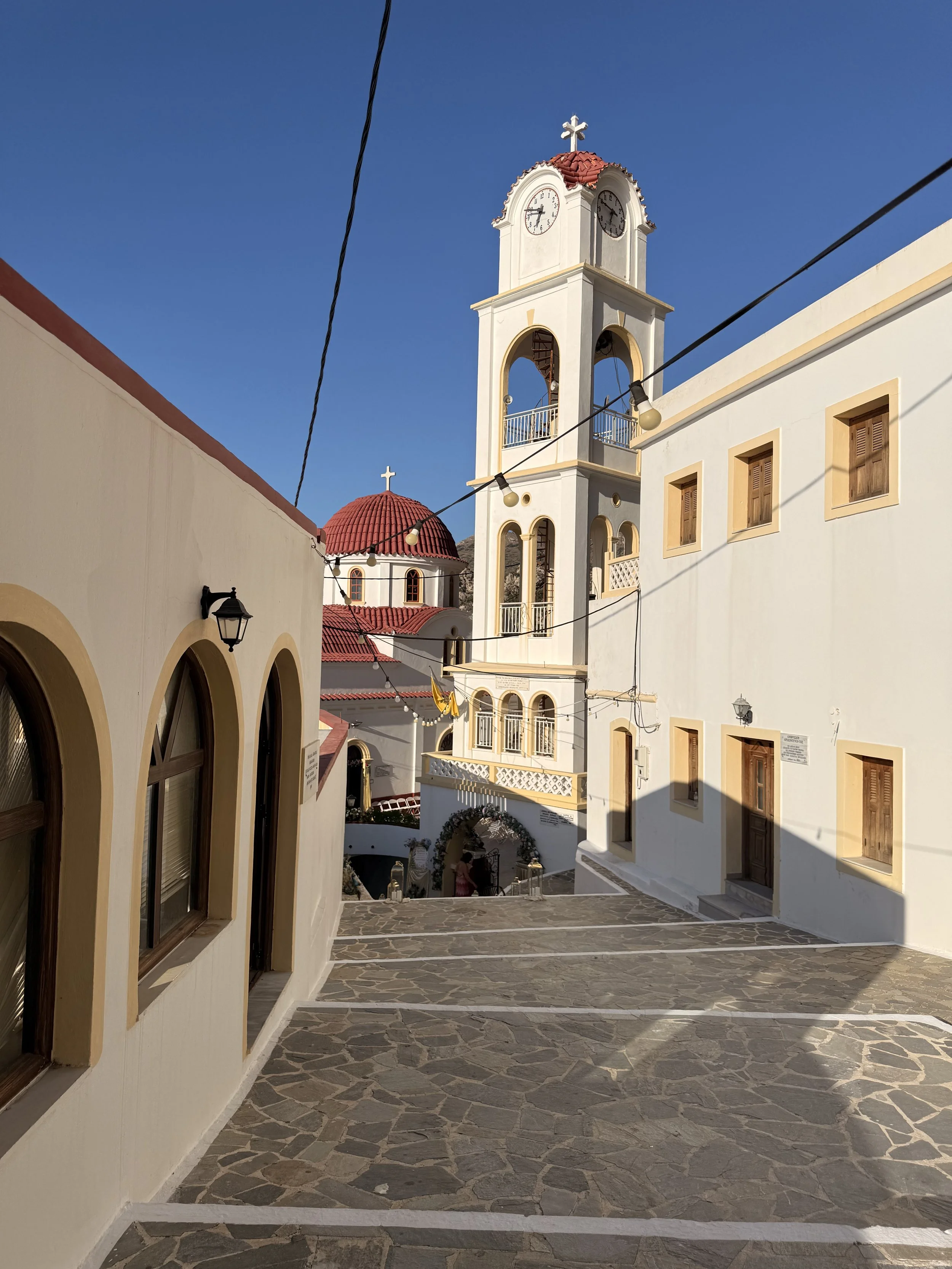 White Greek Orthodox church with clock tower, red tile roofs, and stone-paved courtyard under a clear blue sky