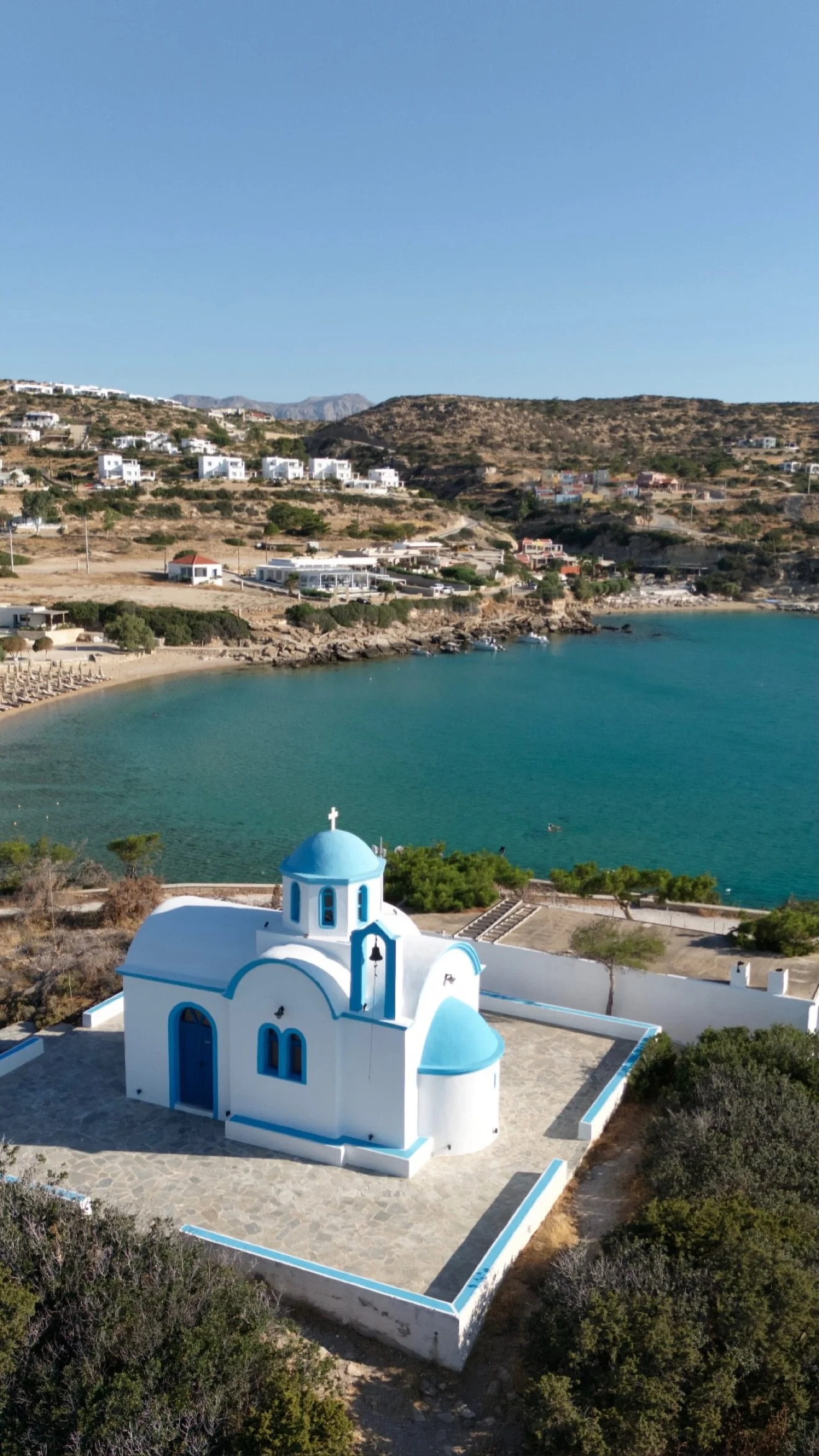 Aerial view of a white-and-blue Greek chapel overlooking a turquoise bay, beach, and hillside village.