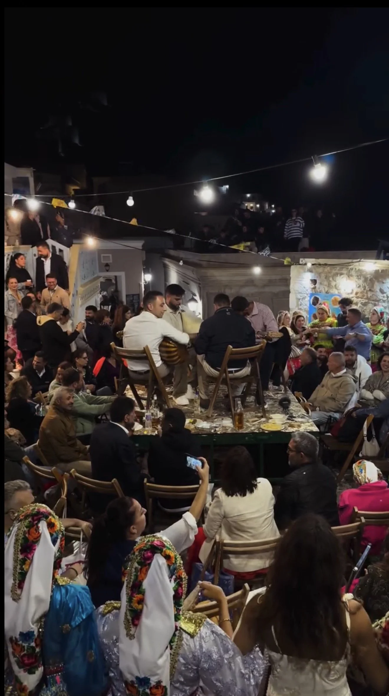 Crowded outdoor nighttime celebration with seated diners, string lights, musicians, and people in colorful traditional costumes.