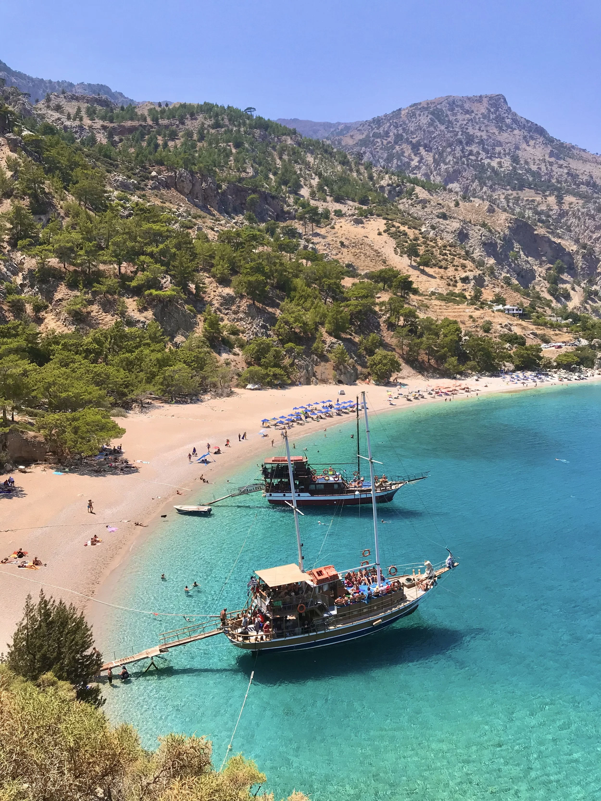 Aerial view of a sandy beach with turquoise water, tour boats, beach umbrellas, and rugged pine-covered mountains.