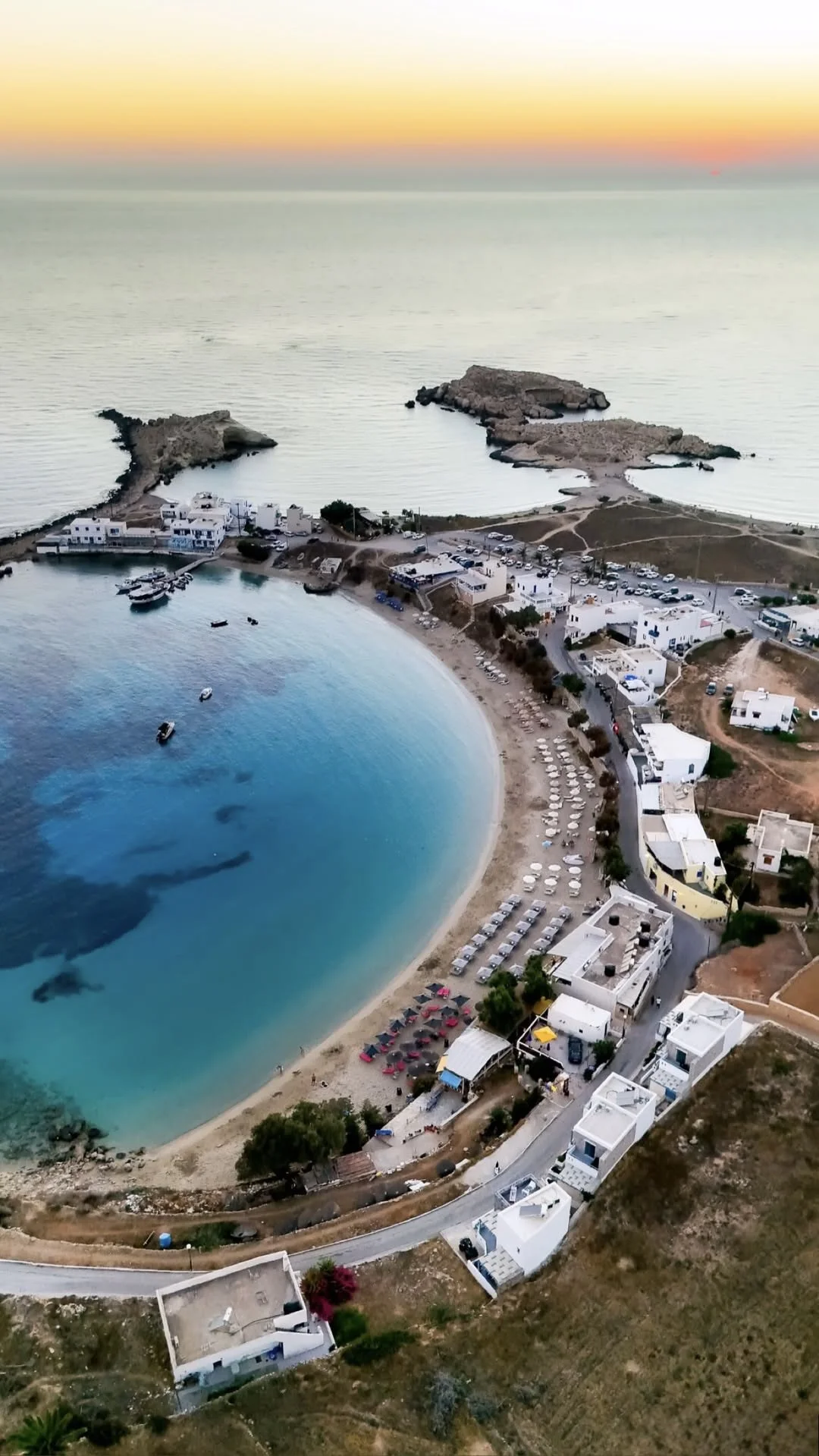 Aerial view of a turquoise beach, whitewashed coastal village, boats, and rocky islets at sunset