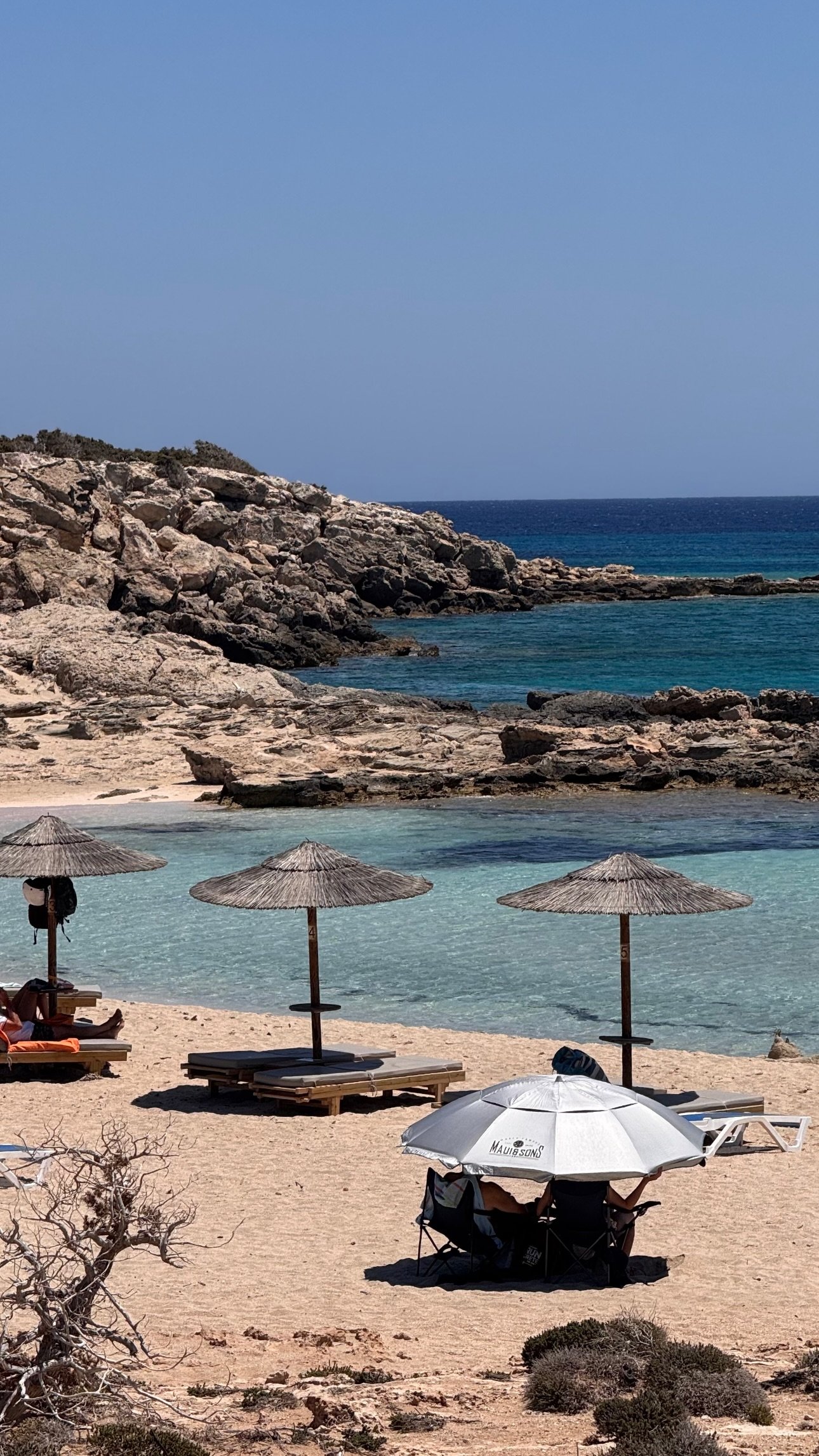 Sunny Mediterranean beach with clear turquoise water, rocky coastline, thatched umbrellas, and lounge chairs.