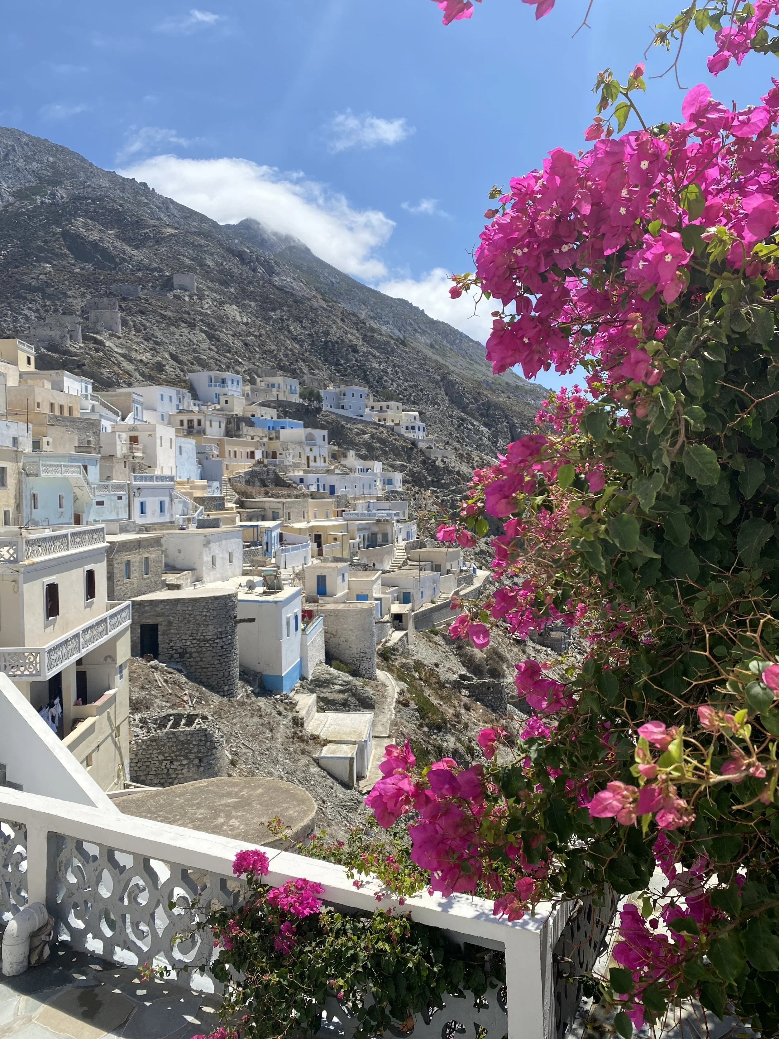 Whitewashed Greek village beneath a rugged mountain, framed by vibrant pink bougainvillea under a blue sky