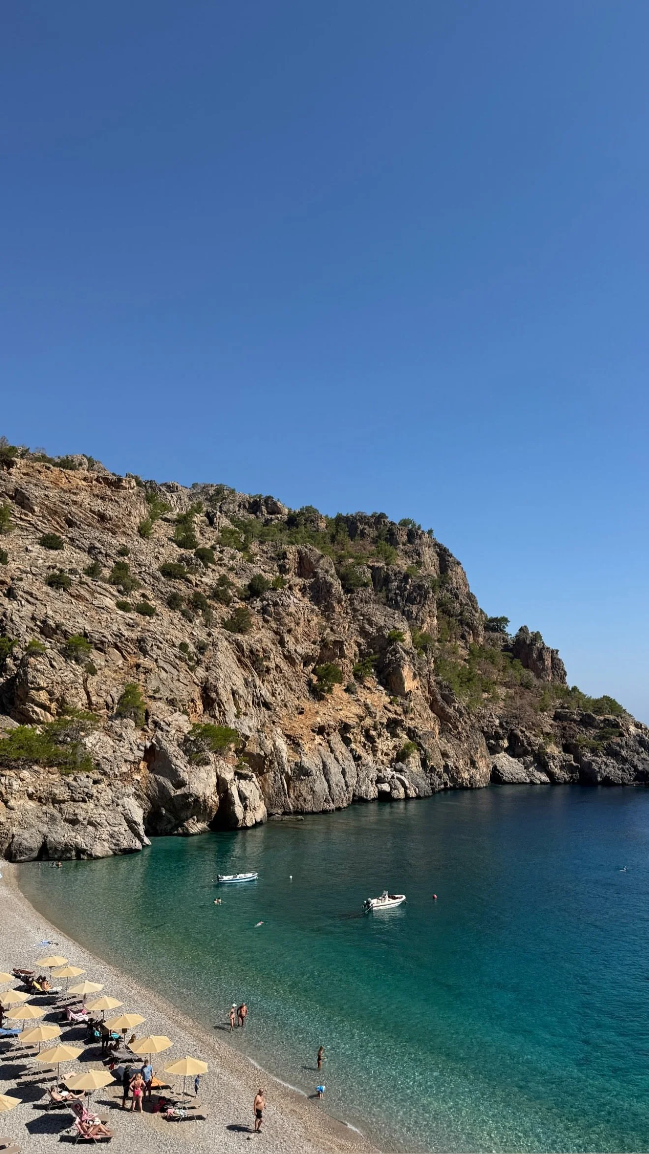 Rocky coastal cove with turquoise water, boats, swimmers, and a pebble beach lined with umbrellas