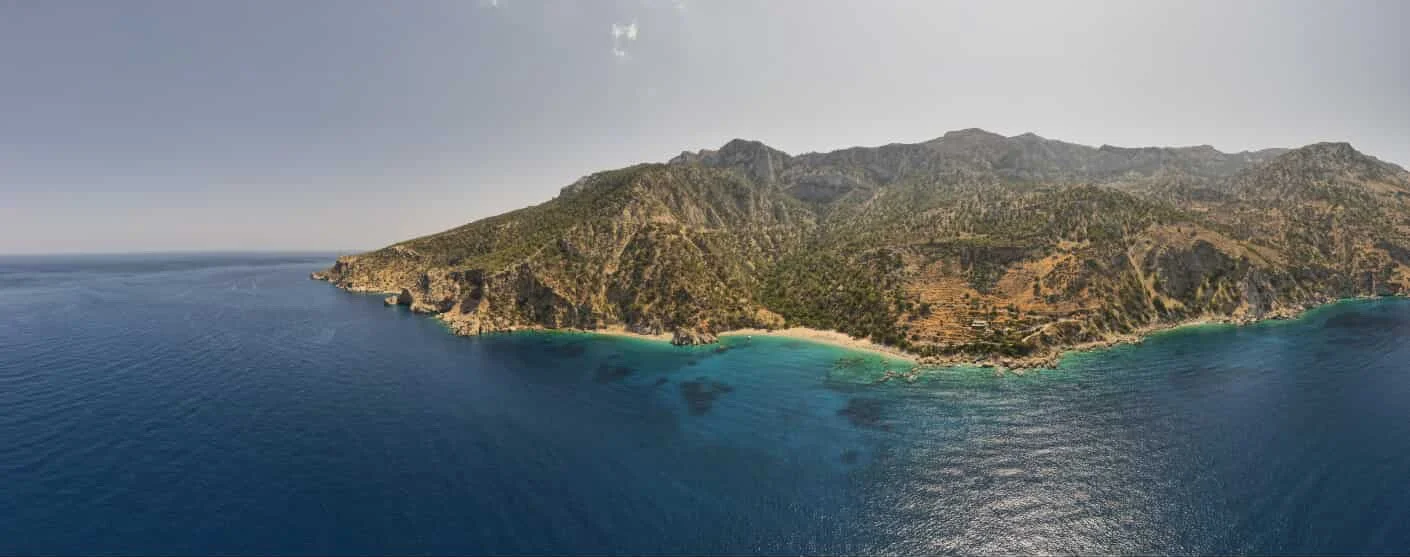 Aerial view of a rugged coastal mountain range and sandy beach beside clear turquoise and deep blue sea