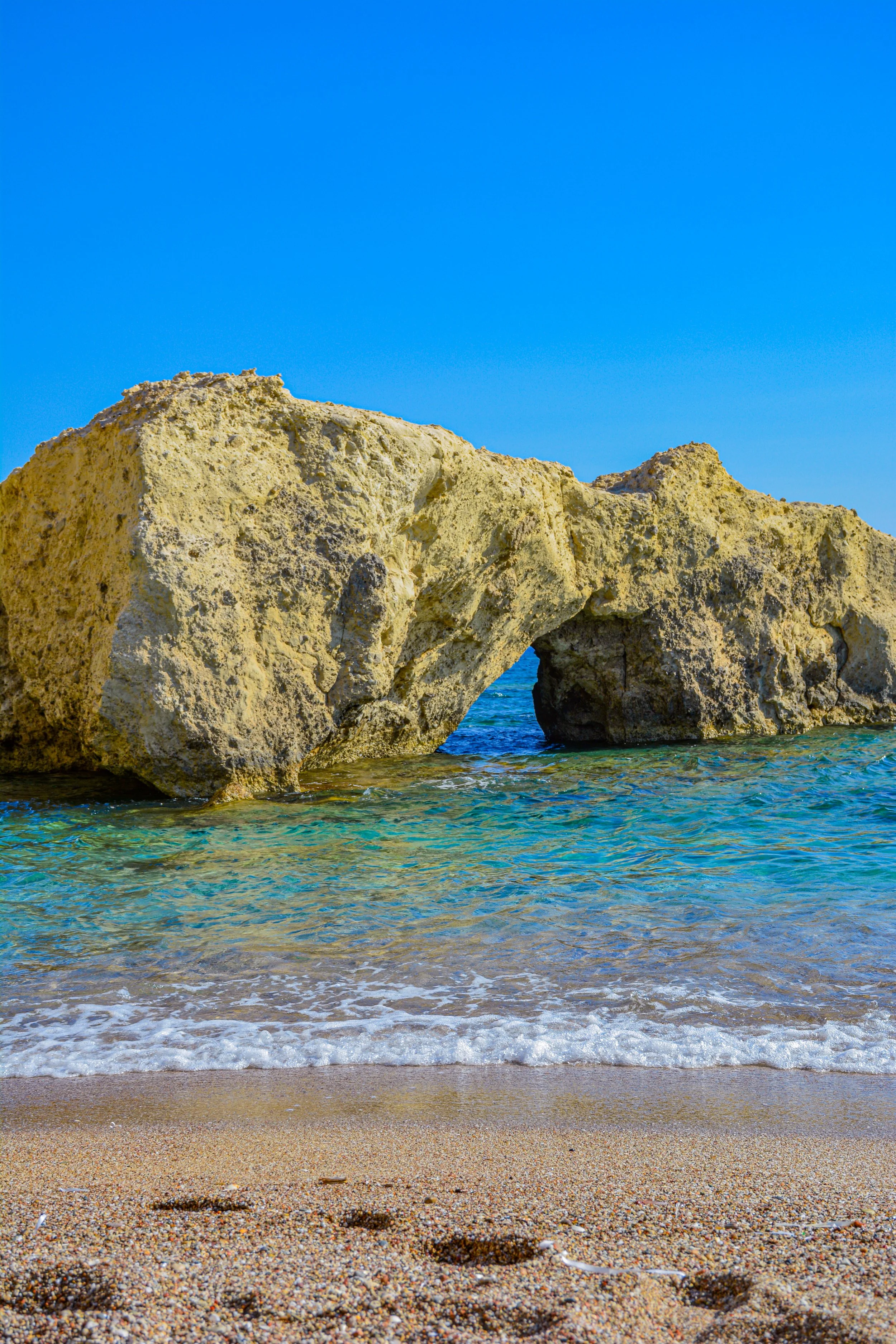 Bright blue sea meeting a sandy beach beneath rugged golden limestone cliffs with a natural rock arch