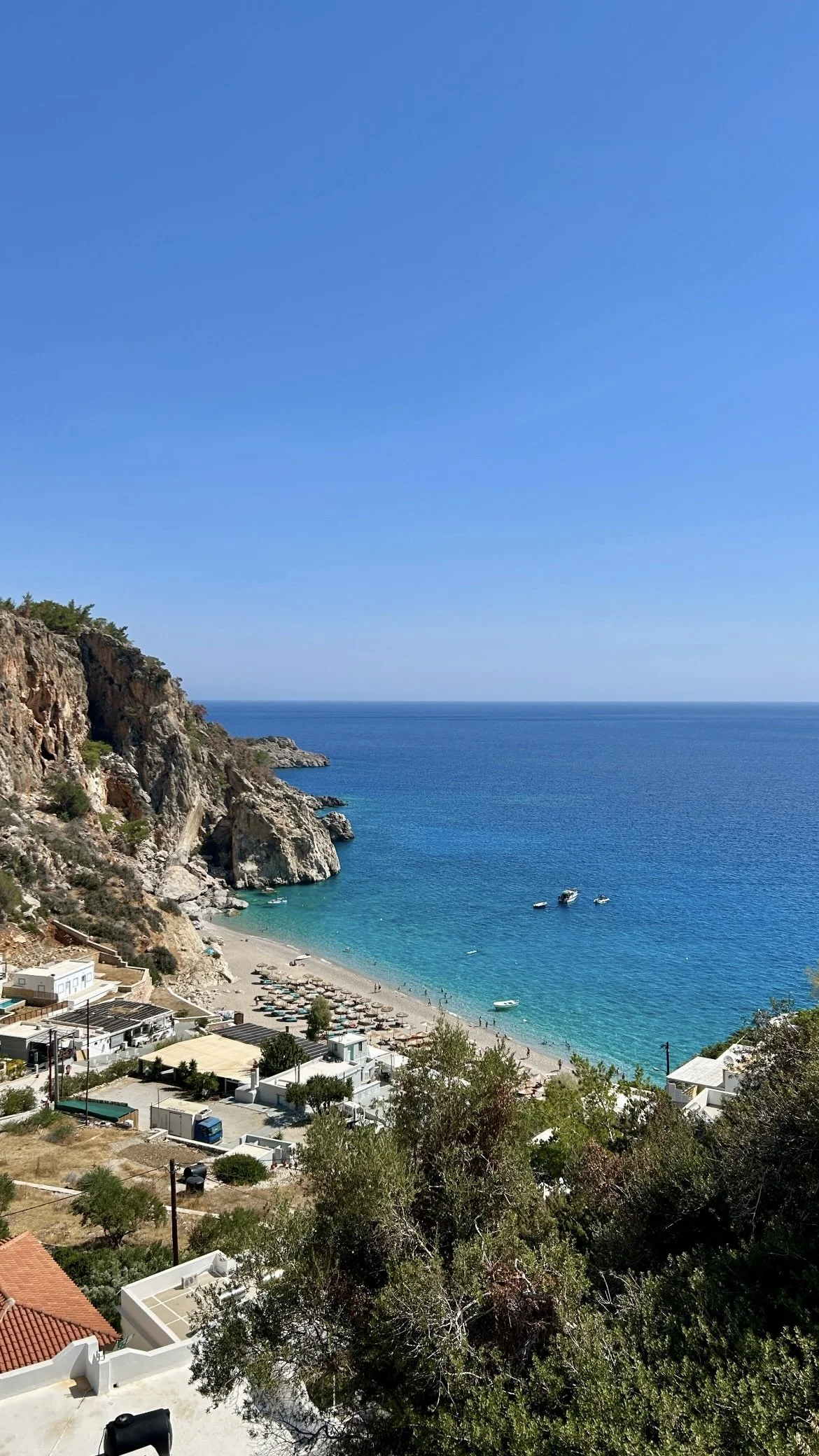Aerial view of a rocky Mediterranean cove with turquoise water, beach umbrellas, boats, and hillside buildings