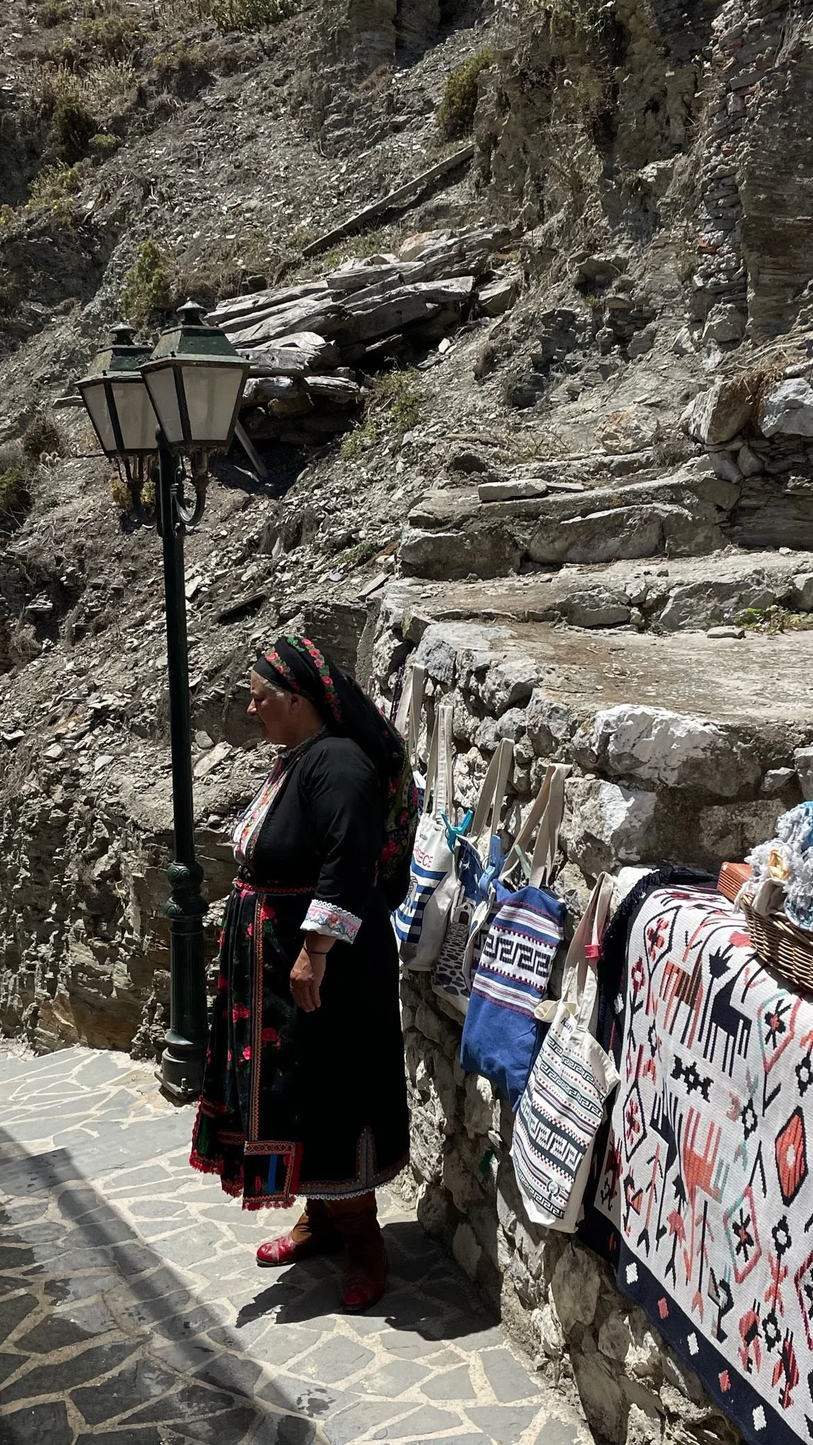 Woman in traditional embroidered clothing beside a vintage streetlamp and colorful woven textiles on a rocky mountain walkway