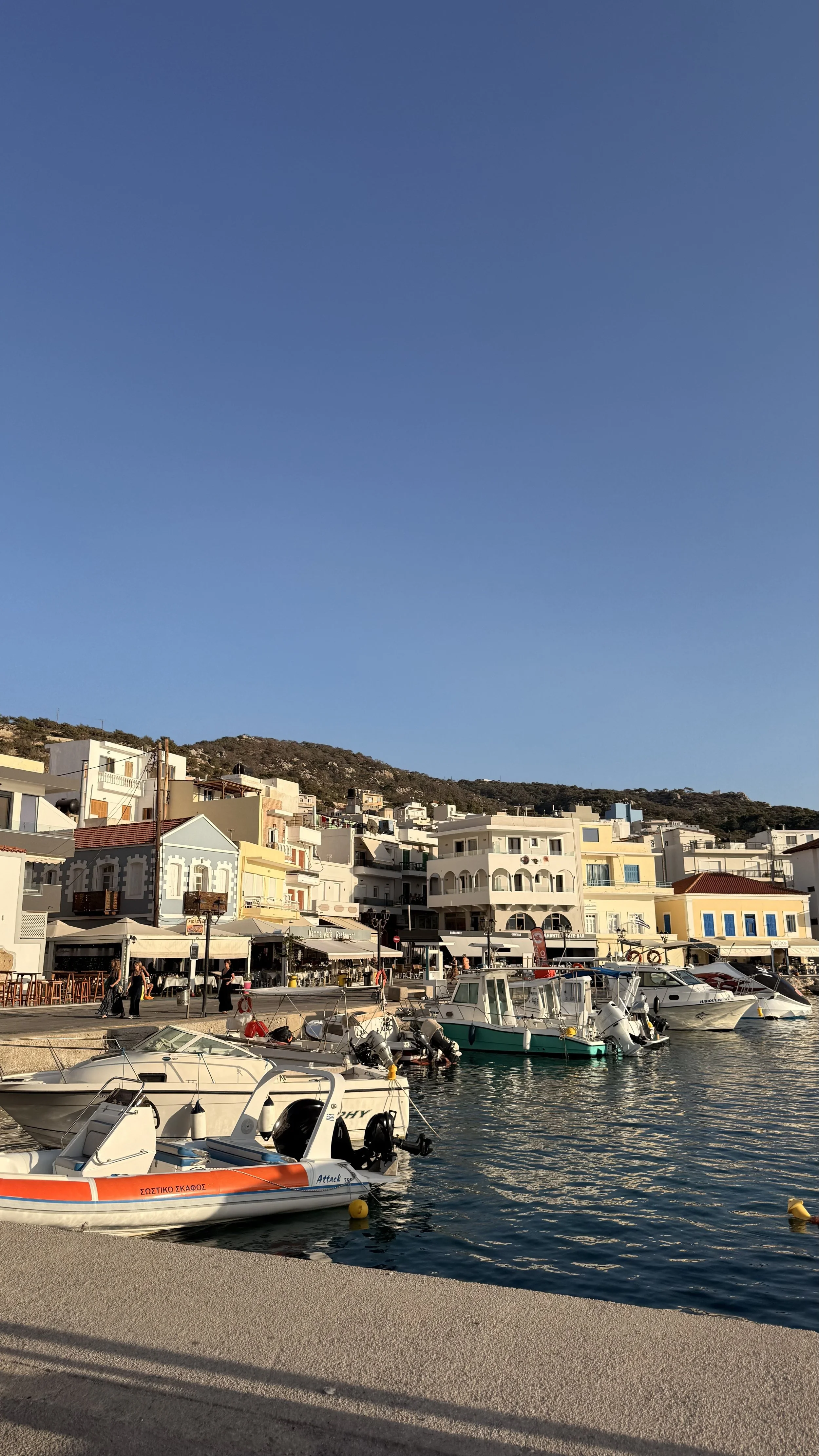 Greek harbor waterfront with fishing boats, seaside restaurants, colorful buildings, and a rocky hillside under a clear blue sky