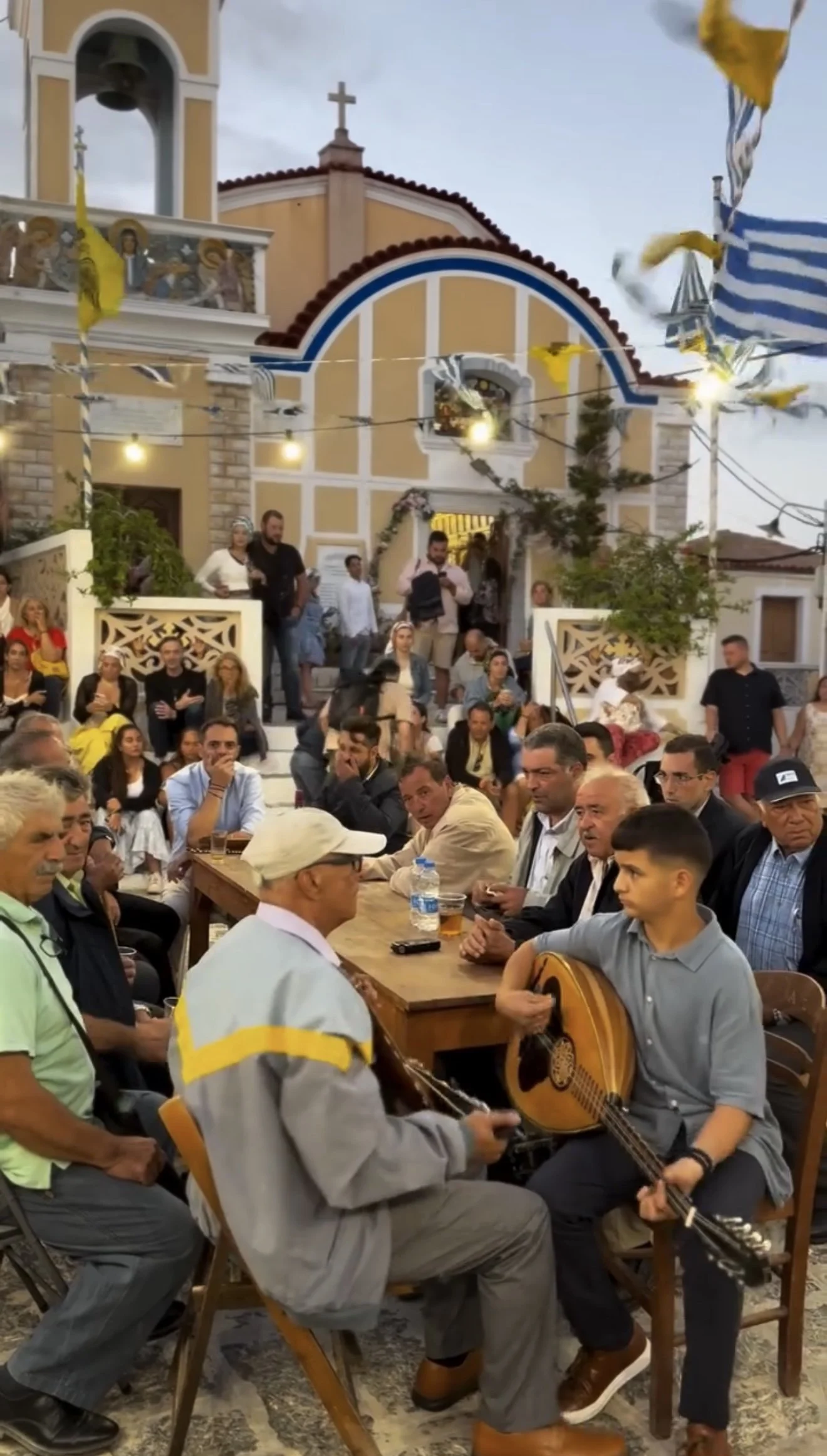 Crowd gathered outside a Greek Orthodox church as musicians play traditional instruments beneath Greek flags and festive decorations.