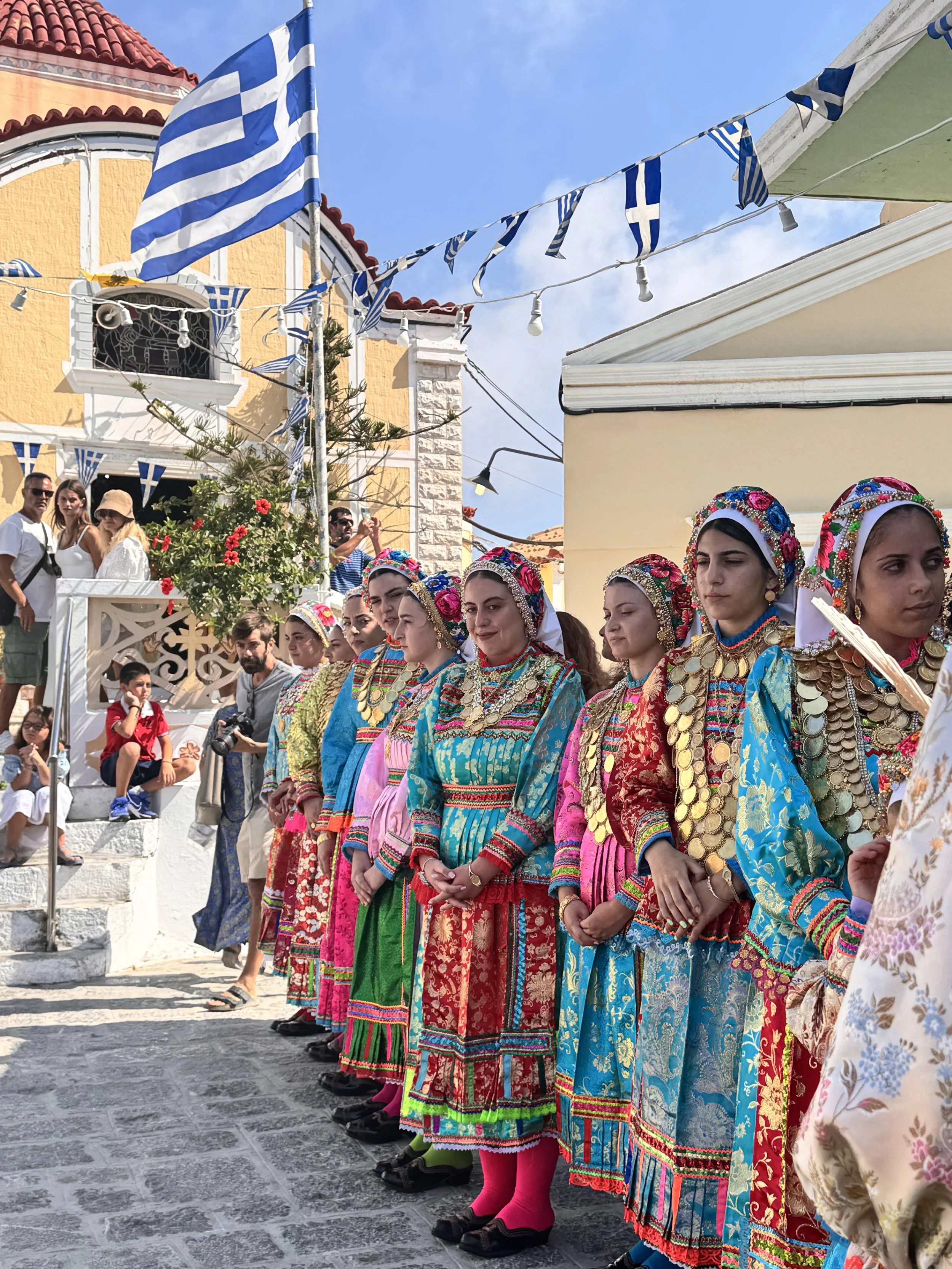 Women in colorful traditional Greek costumes stand during a festival beneath Greek flags and string lights.