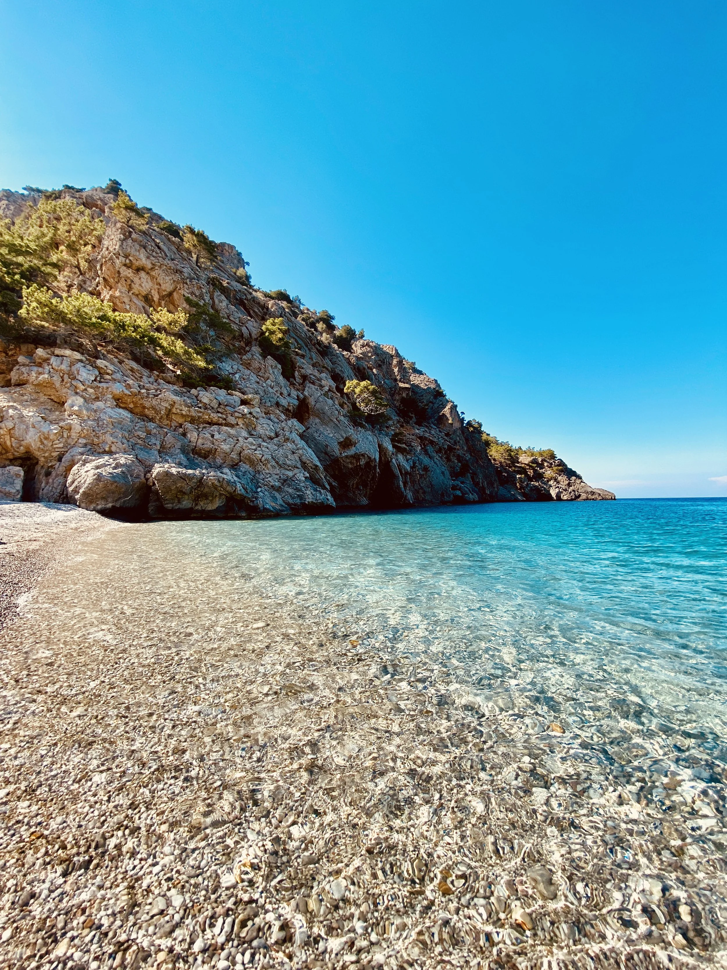 Rocky Mediterranean coastline with clear turquoise water, pebbled beach, and bright blue sky