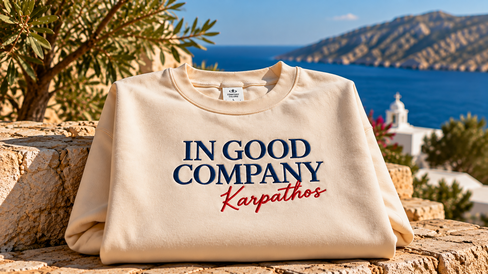 Cream sweatshirt reading “In Good Company Karpathos” displayed on a stone terrace overlooking the blue sea and mountains of Karpathos, Greece.