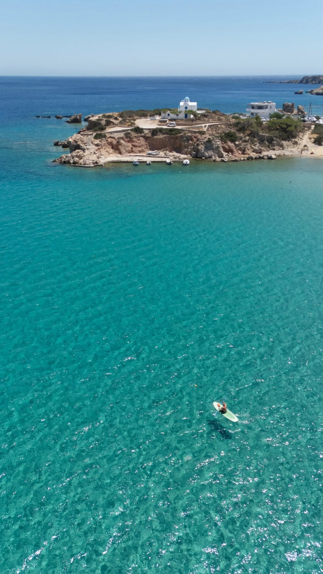Aerial view of a rocky Mediterranean coastline with a white domed chapel, turquoise water, small boats, and a paddleboarder.