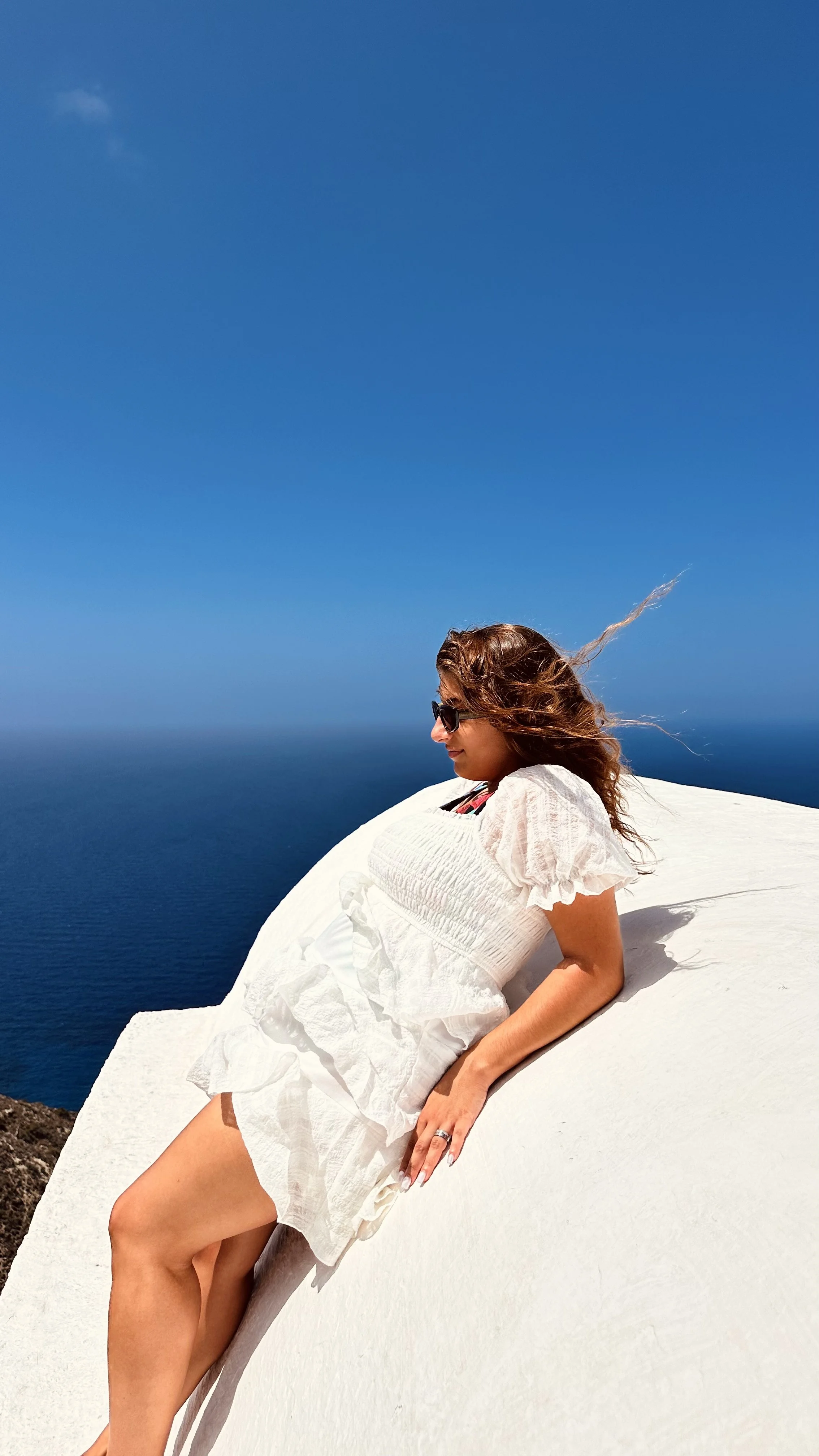 Woman in sunglasses and a white dress sitting on a boat against a bright blue sea and sky
