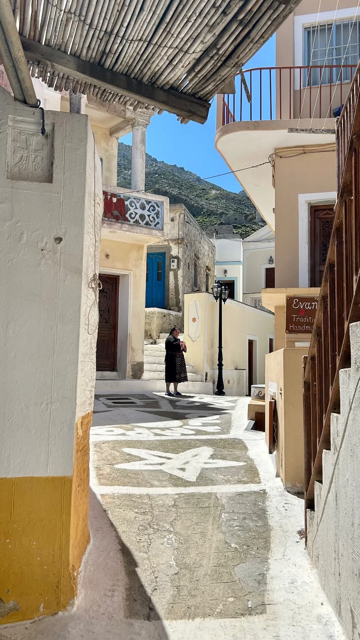 Narrow whitewashed village alley with traditional homes, a woman, Greek-style lamp, mountain backdrop, and handmade crafts shop