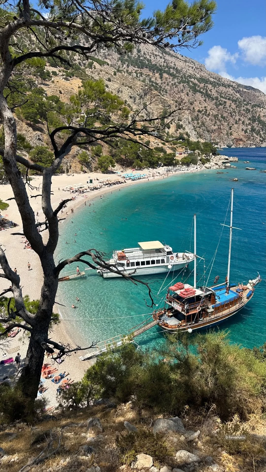 Aerial view of a turquoise Mediterranean cove with a pebble beach, swimmers, tour boats, sailboats, pine trees, and rugged mountains.