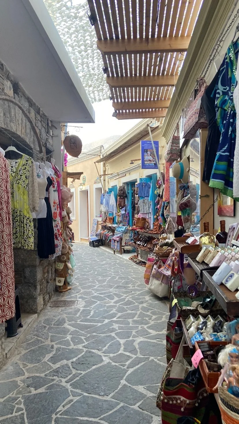 Narrow stone-paved shopping alley lined with clothing, accessories, souvenirs, and local crafts under a wooden slatted canopy