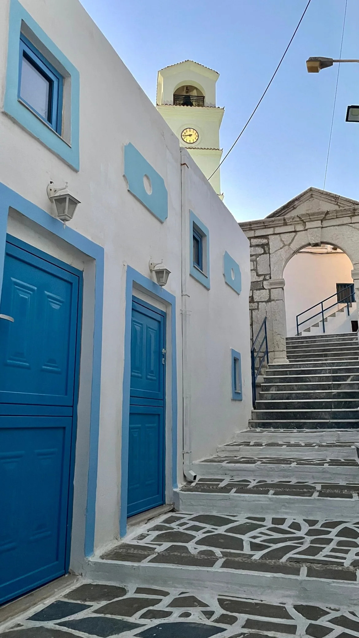 Whitewashed Greek island alley with bright blue doors, stone paving, an arched gateway, and a clock tower under a clear blue sky