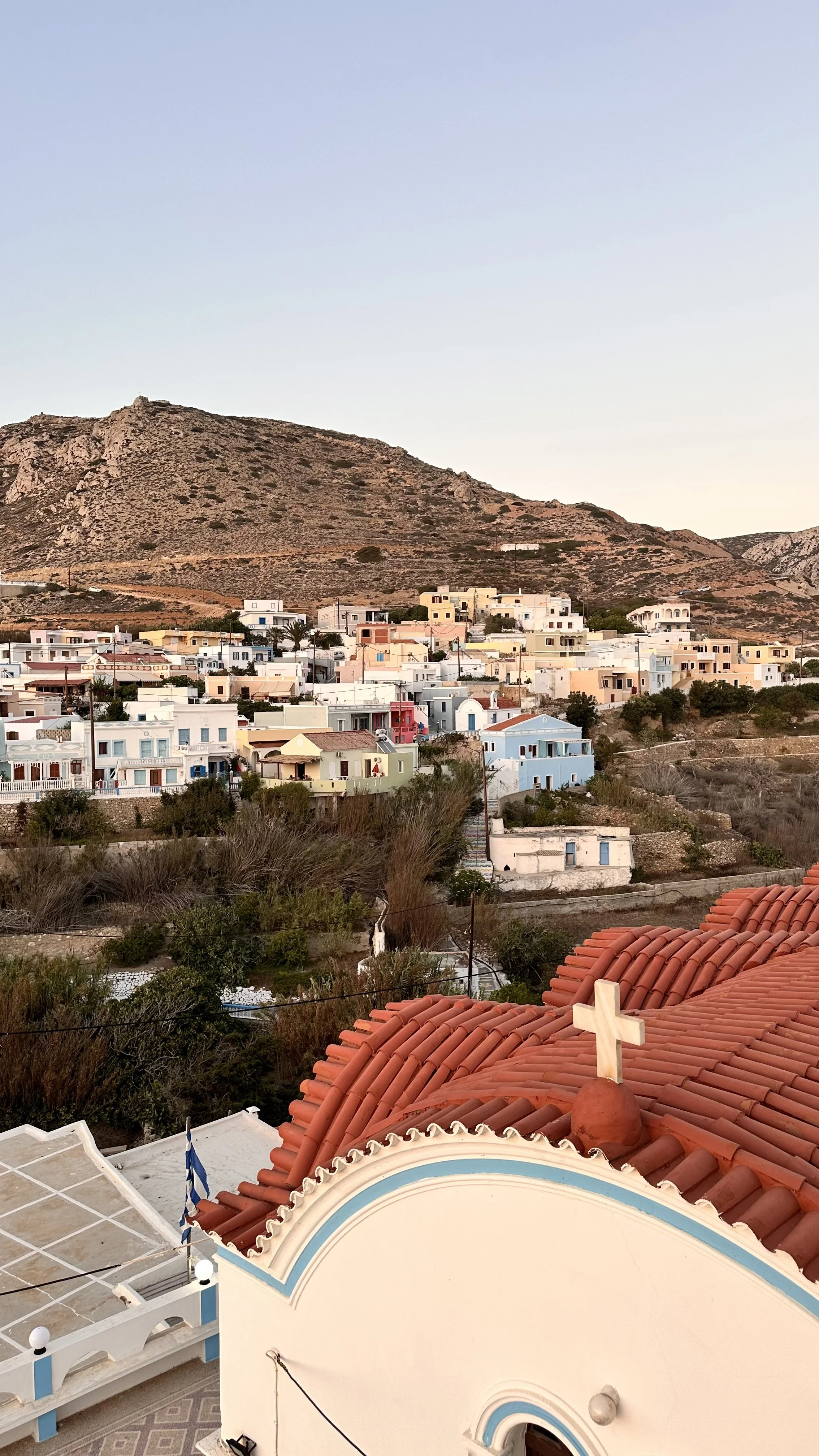 Colorful Greek village beneath a rugged rocky mountain, with pastel homes, terracotta roofs, and a church in the foreground.