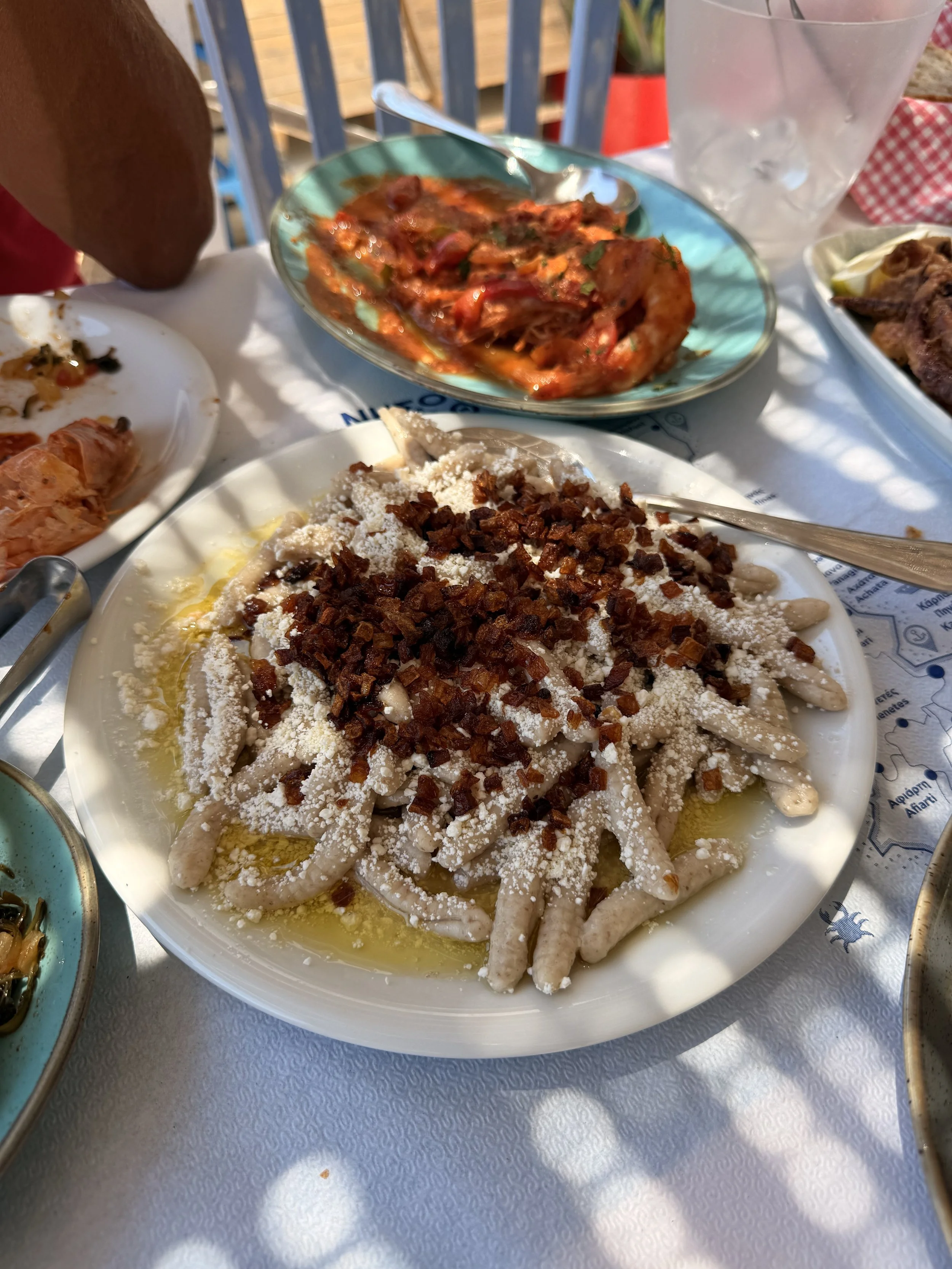 Greek meal featuring a large plate of creamy pasta topped with feta and crispy onions, with tomato salad and grilled meat on the table