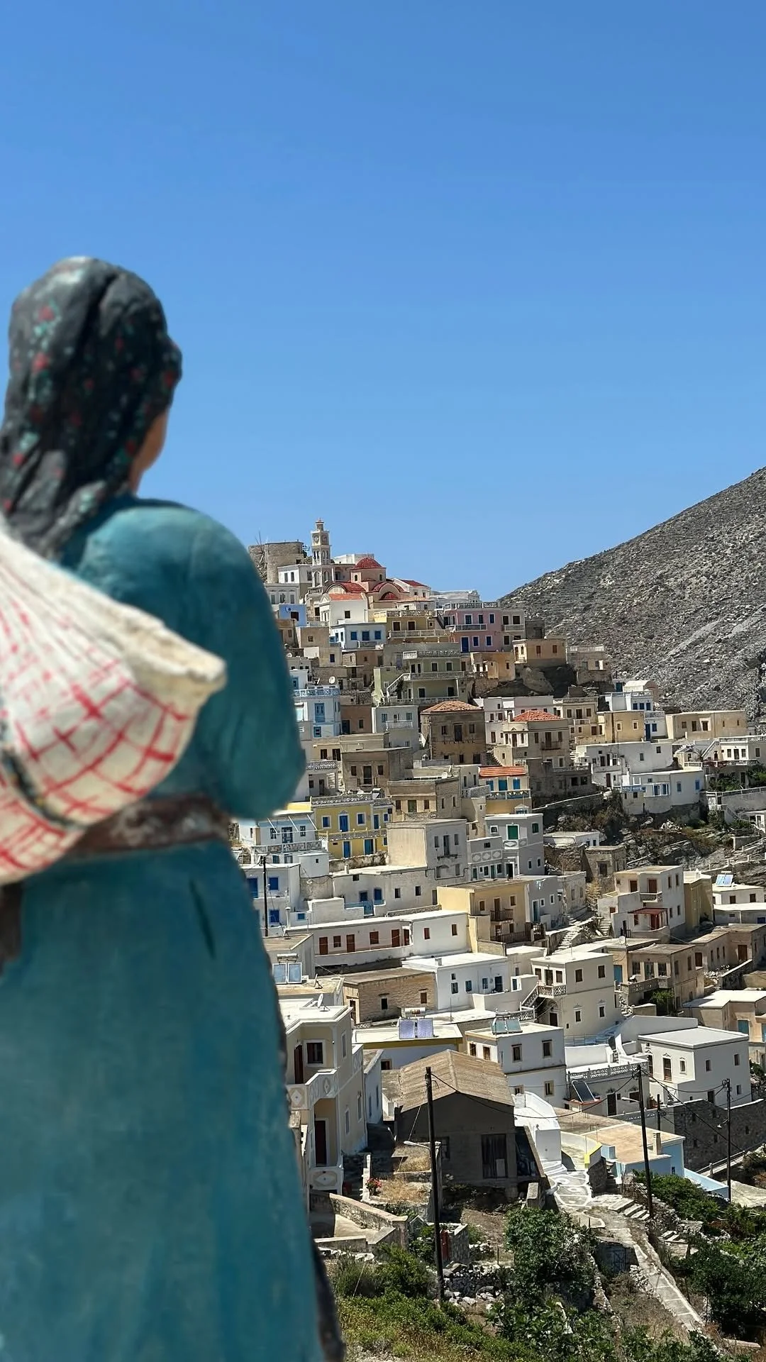 Woman in a patterned headscarf overlooking a colorful hillside village beneath a clear blue sky