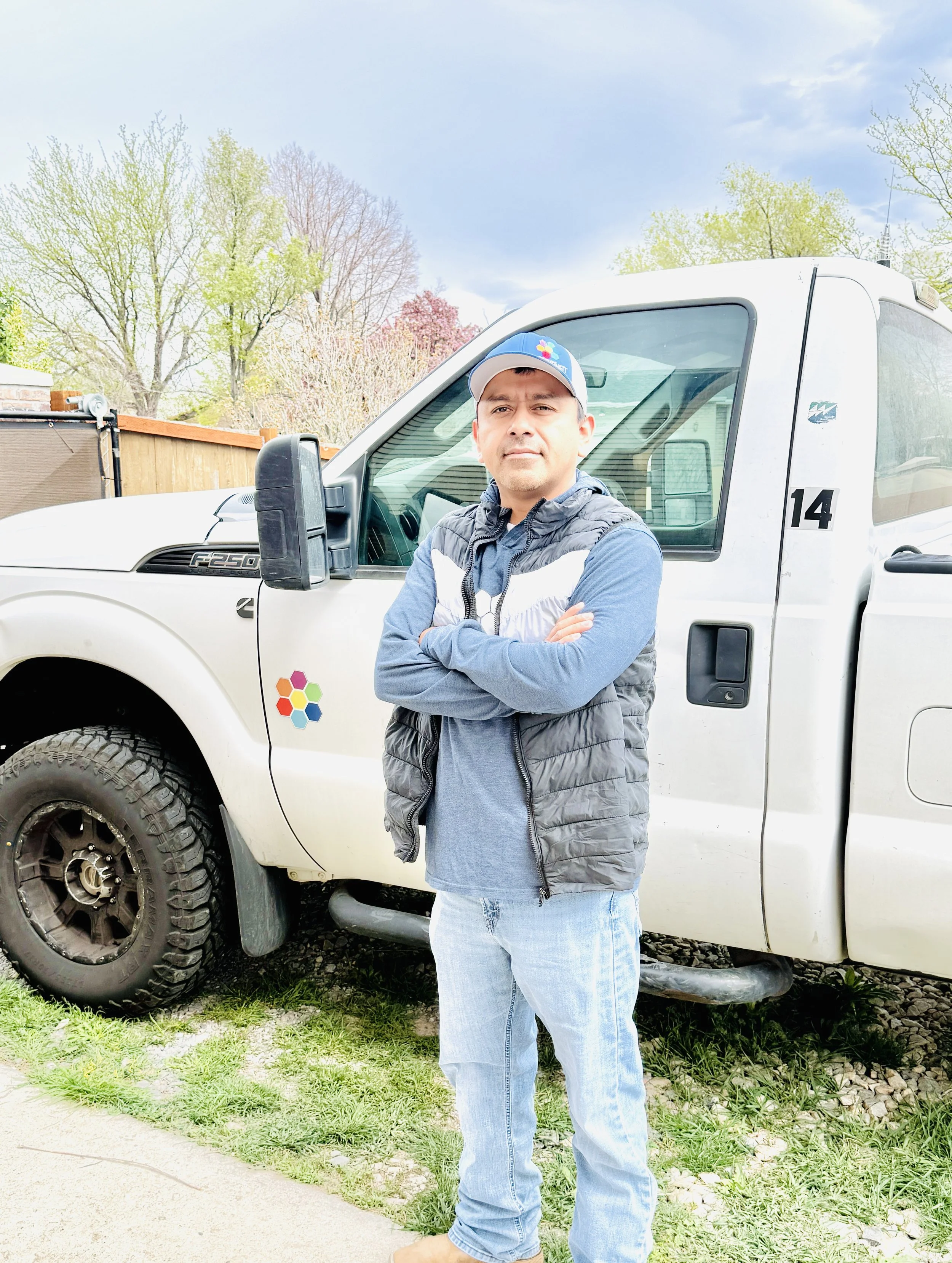 Man in a blue cap and vest standing with crossed arms beside a white Ford F-250 truck outdoors
