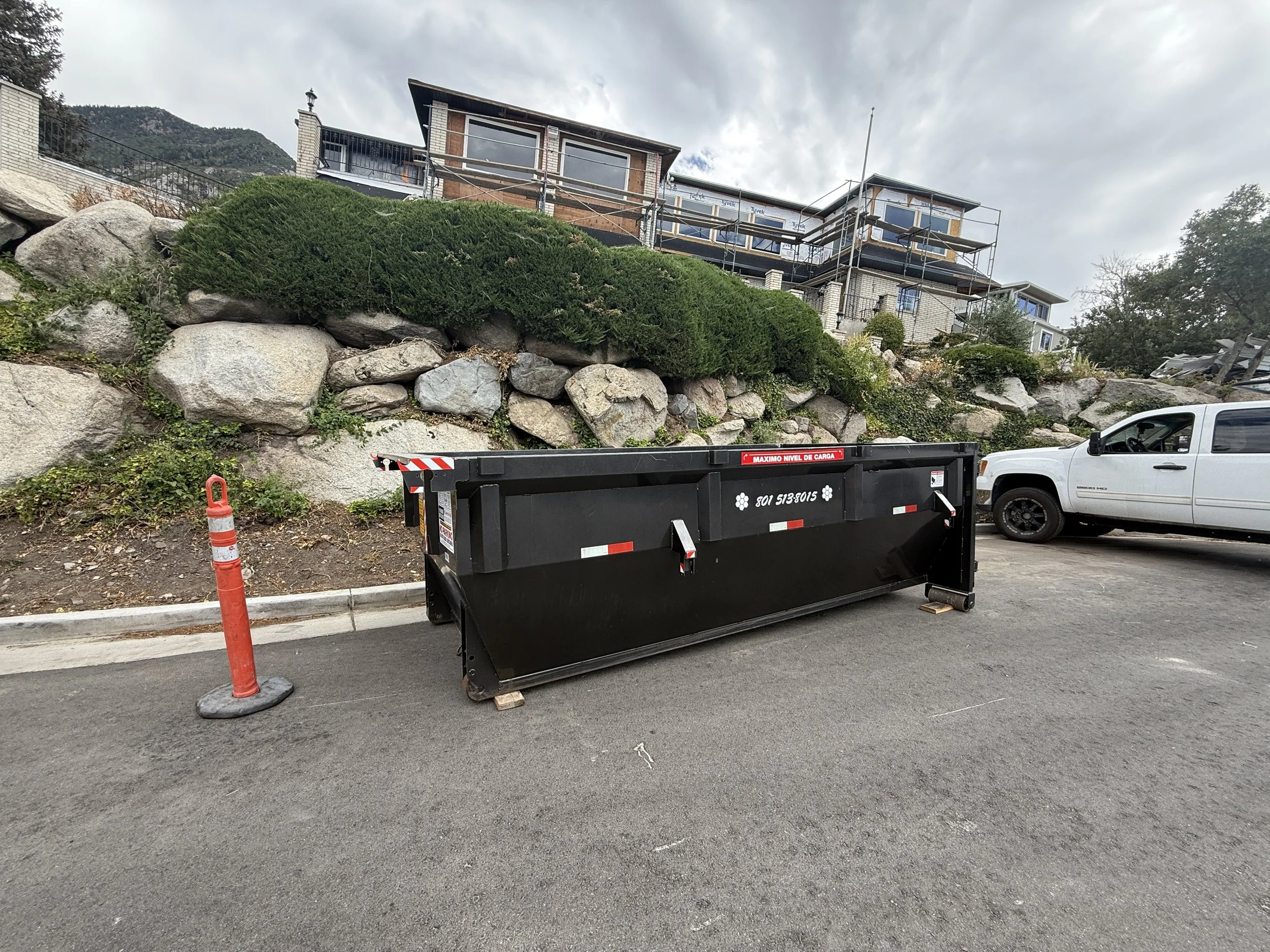 Large black construction dumpster parked outside a partially renovated hillside house with scaffolding, rocky landscaping, and a white pickup truck.