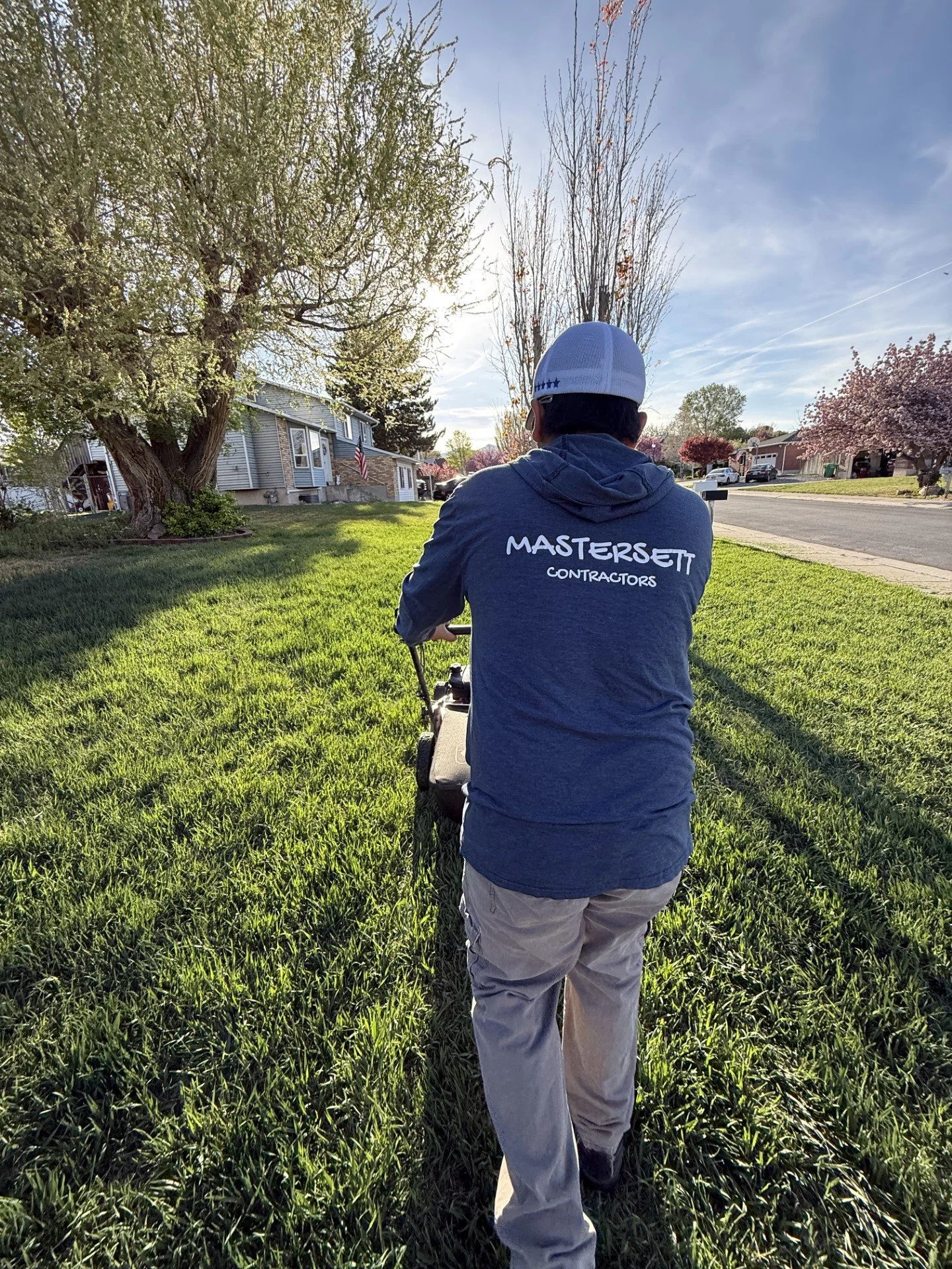 Contractor wearing a MastersElite Contractors shirt mowing a suburban lawn on a sunny day.