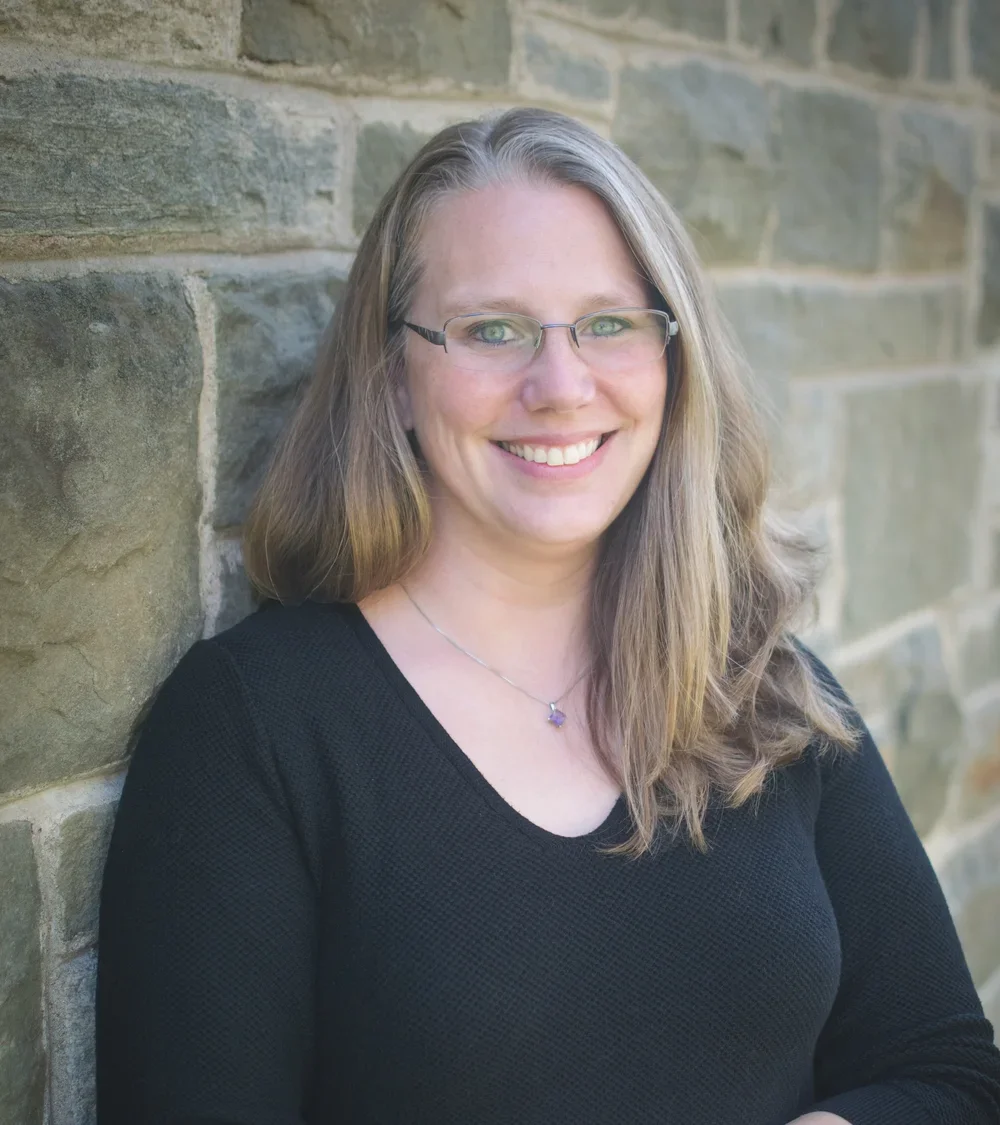 a woman with light brown hair and a black shirt standing in front of a brick wall