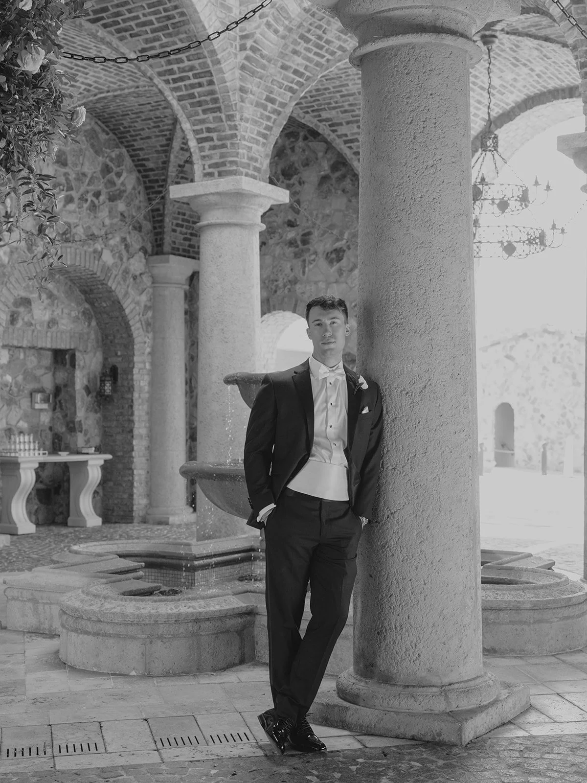 Groom in a tuxedo posing beside a fountain in a historic stone courtyard